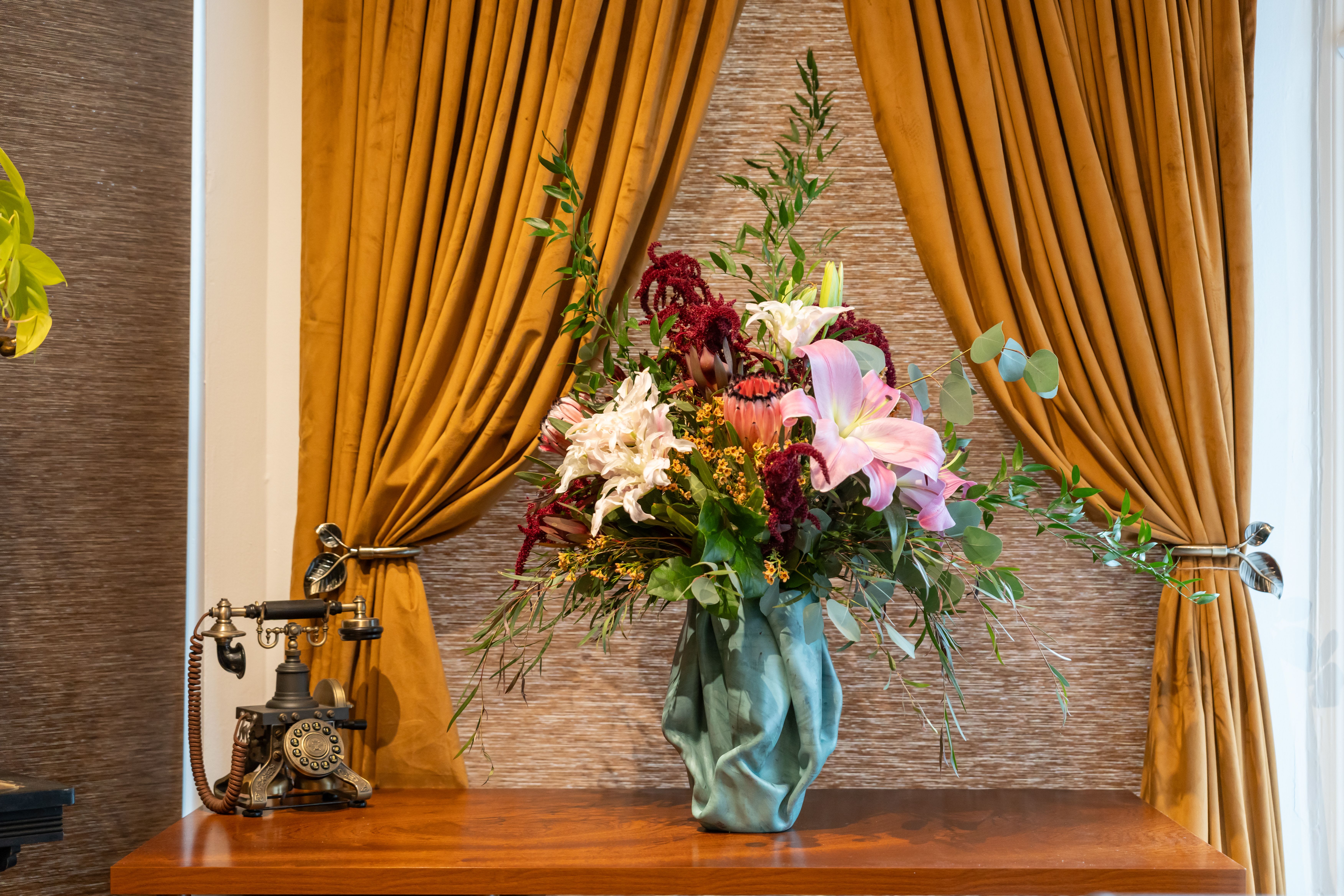 Glamour Tramp - Glamour Tramp—because who says high maintenance can’t also be high style? This over-the-top arrangement struts its stuff with fiery proteas, bold roses, sassy snapdragons, and enough lush greenery to make a jungle jealous. But the real star of the show? The Mega Leather Vase, exclusively designed for Chartreuse by Roje by local leather artisan Ian James. Hardened leather, impeccable craftsmanship, and a vibe that says, “Yes, I’m extra”—this vase is the perfect partner for a bouquet that’s equal parts glam and grit. Glamour Tramp: where elegance meets a little edge.  Want her in a different vase? Call us at 415.374.7812