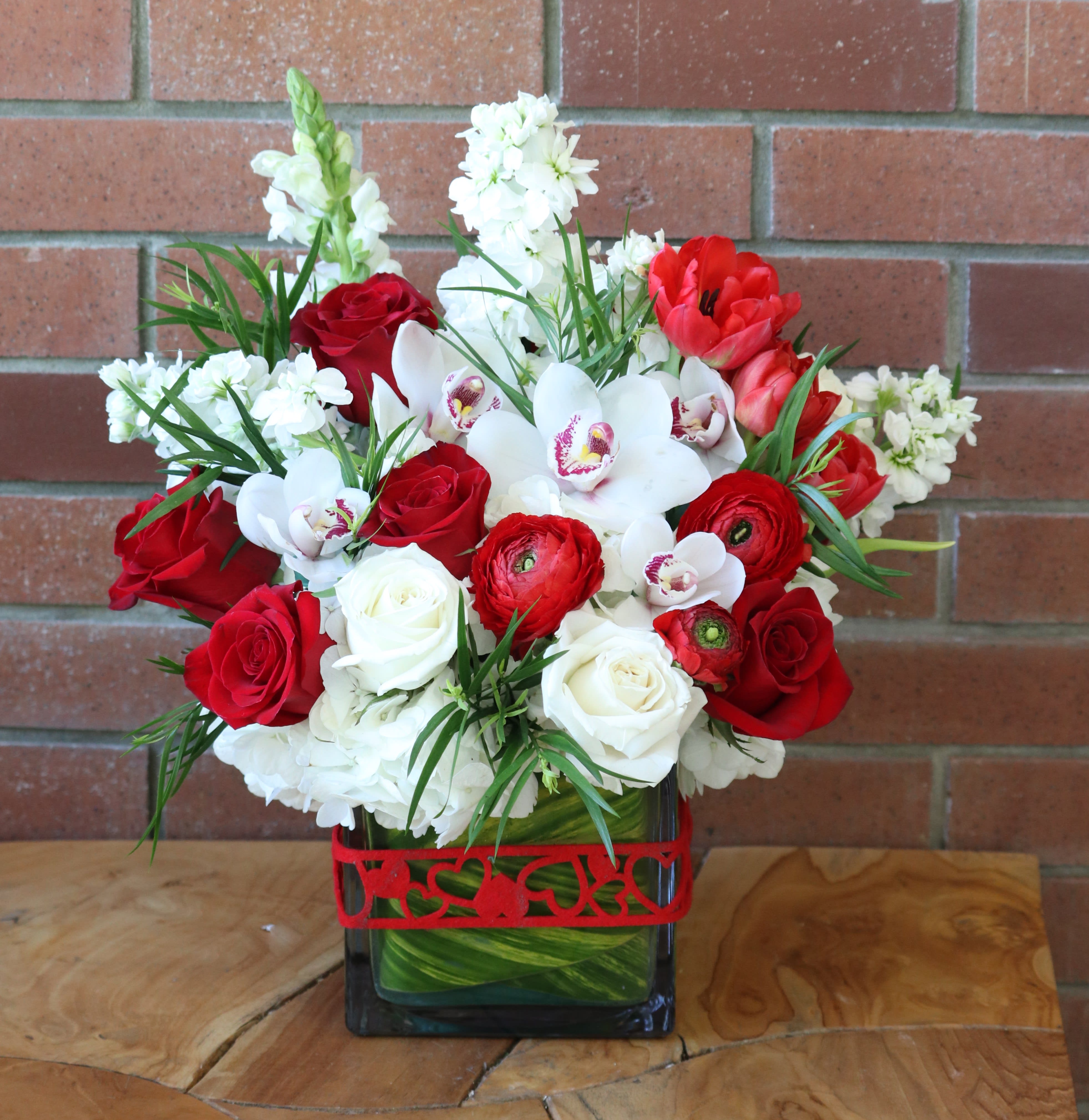 Bright In Reds - Lush floral arrangement in reds and whites. Red ranunculus, red roses, orchids, hydrangeas, stock, snap dragons and lush greenery in a large cube with a green leaf and ribbon.