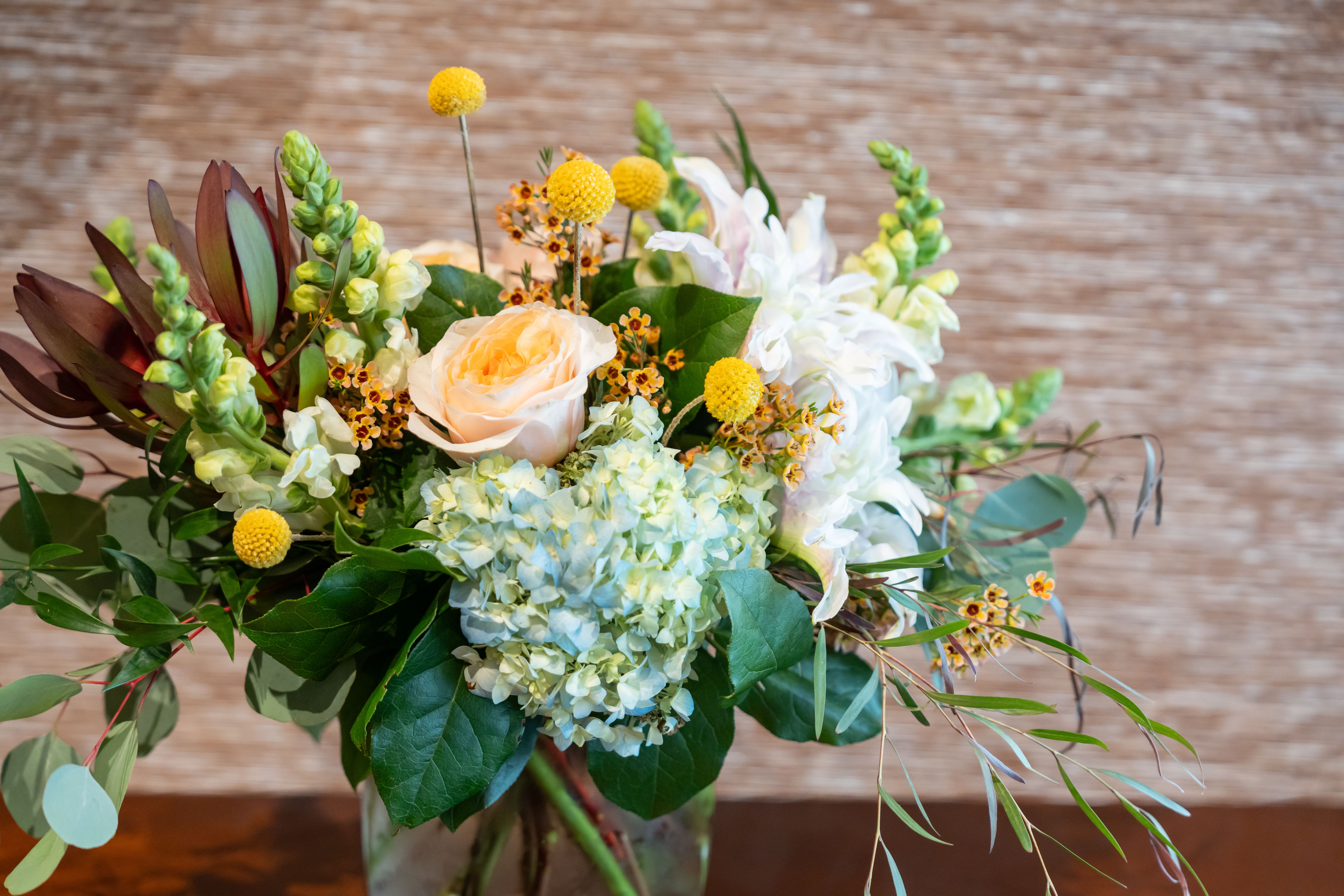 Morning Tea - Darling, let us introduce you to the most delightful way to start your day. This utterly chic arrangement is a perfect marriage of soft and sassy, featuring a show-stopping pale blue hydrangea that simply demands attention. A perfectly peachy garden rose adds that &quot;just woke up like this&quot; elegance, while perky yellow craspedia balls (we like to call them nature's sunshine drops) bring the morning energy we all crave.  We've added pristine white snapdragons because, honey, every arrangement needs a little drama, and deep burgundy leucadendron leaves for that touch of sophistication. The golden waxflower sprays are giving us that effortless &quot;I didn't try too hard&quot; vibe, while eucalyptus and mixed greenery keep it fresh and modern.  Artfully arranged in a sleek glass vase, this beauty is serving breakfast-at-Tiffany's realness with a modern twist. It's the perfect &quot;treat yourself&quot; moment or &quot;I'm sorry I forgot our anniversary&quot; gesture (we don't judge).   Care Tips for Your Morning Moment: Keep the water fresh and cool (like your attitude) Avoid direct sunlight (it's already bright enough) Remove fallen leaves (nobody likes a messy moment) Enjoy 5-7 days of pure floral fabulousness