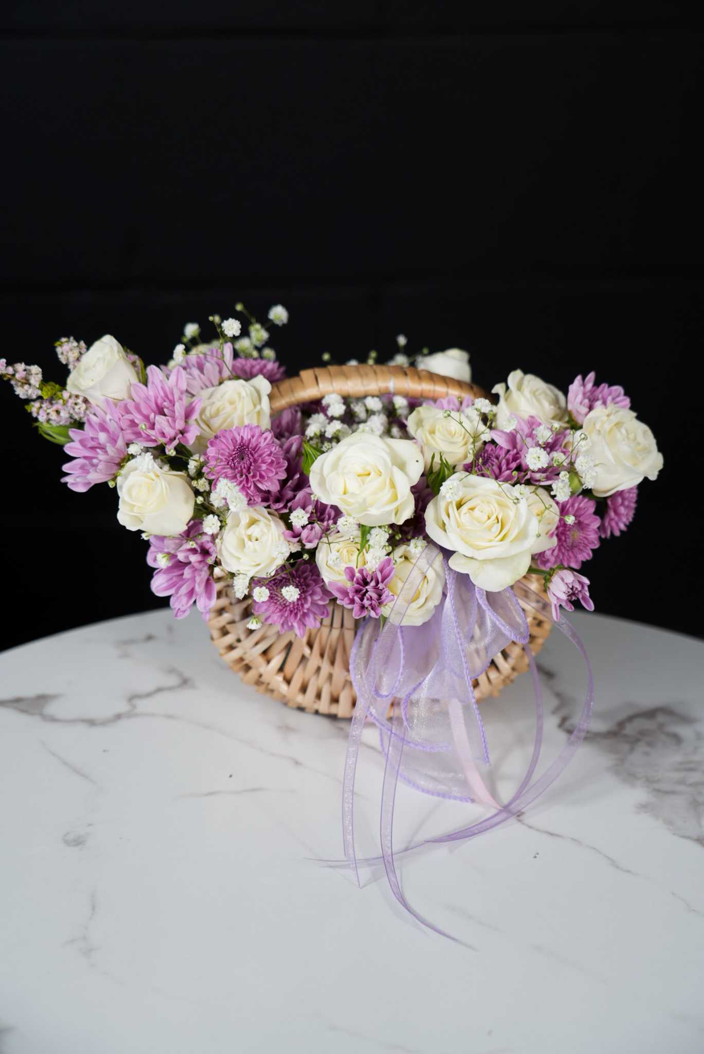 Flonet Petit Lavender Dreams Basket  - The Lavender Dreams Basket is an elegant and serene arrangement that beautifully combines the classic beauty of white roses with the charming allure of lavender chrysanthemums. Nestled in a delicate wicker basket, this arrangement is perfect for expressing heartfelt sentiments and bringing a touch of grace to any occasion.  The soft white roses symbolize purity and elegance, while the lavender chrysanthemums add a touch of whimsy and charm. Accents of baby's breath and other delicate white flowers enhance the overall aesthetic, creating a harmonious blend of colors and textures. The basket is adorned with a lavender ribbon, adding a finishing touch of sophistication.  Dimensions:  Basket diameter: 10 inches Approximate height: 12 inches Care Instructions:  Keep the flowers in a cool, dry place, away from direct sunlight. Add a small amount of water daily to keep the flowers fresh. The Lavender Dreams Basket is an ideal gift for celebrations, anniversaries, or simply to brighten someone's day with its timeless beauty and soothing colors.