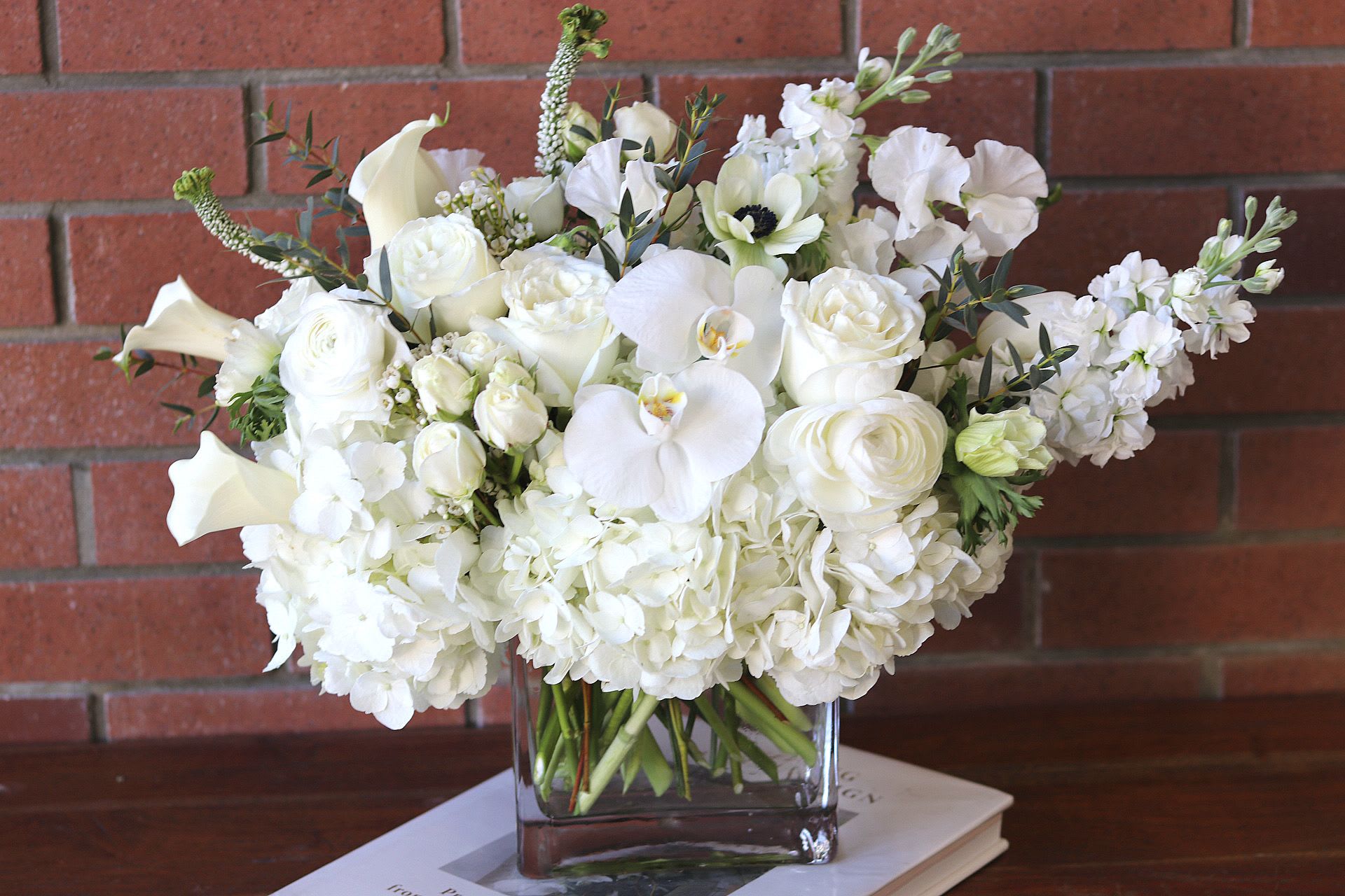 Pure Elegance  - The &quot;Pure Elegance&quot; bouquet is a breathtaking all-white arrangement that embodies sophistication and grace. Designed in a modern rectangular clear glass vase, this stunning piece showcases a harmonious blend of luxurious blooms, creating a serene and captivating display.  At the foundation, voluminous white hydrangeas provide a lush, cloud-like texture, serving as an exquisite backdrop for the other delicate flowers. Nestled among them, the elegant Playa Blanca roses exude a soft, romantic charm, their pristine petals adding depth and richness to the arrangement.  Delicate white anemones peek out with their striking dark centers, offering a touch of intrigue and contrast, while fragrant white stock infuses the bouquet with a delightful scent, enhancing the overall sensory experience. Completing this elegant composition are the enchanting white sweet peas, known for their delicate fragrance and sweet appearance, which add a whimsical touch.  At the forefront, the stunning Phalaenopsis white orchid blooms stand  proud, ranunculus, spray roses and white mini calla lilies showcasing their graceful lines and luxurious texture. Their presence elevates the arrangement, making it a true statement piece.  The &quot;Pure Elegance&quot; bouquet is perfect for any occasion, from weddings to sophisticated events, or as a chic centerpiece in a modern home. Its all-white palette radiates tranquility and class, making it a timeless choice for those seeking understated beauty.