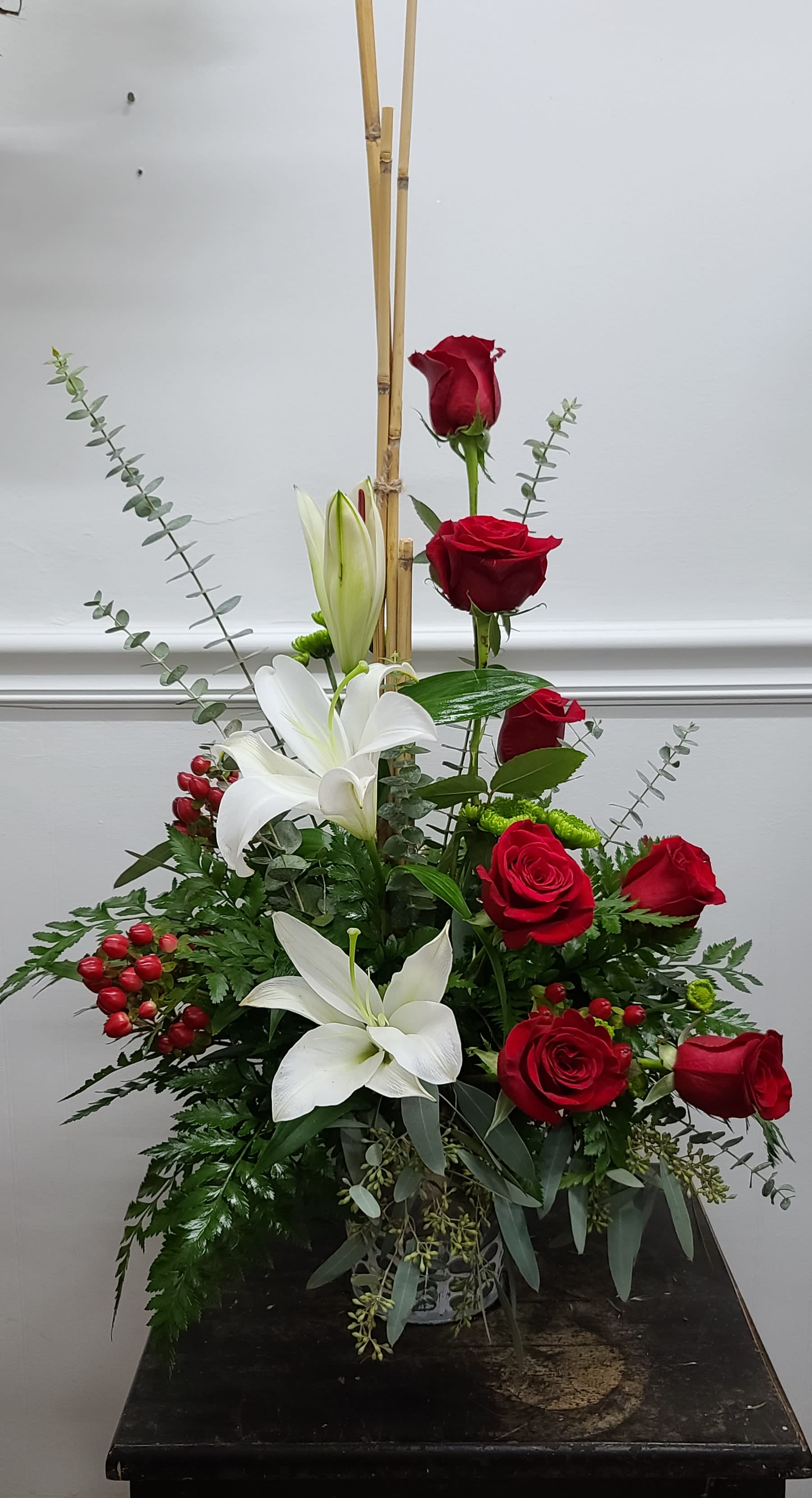 Winter Roses (Shown Deluxe) - White Lilies, Hypericum berries, and Red Roses in a traditional display, accented with bamboo  and eucalyptus. This arrangement is a gorgeous display of love and fond memories. This arrangement looks stunning in an entryway, dining table or kitchen island for the season. 