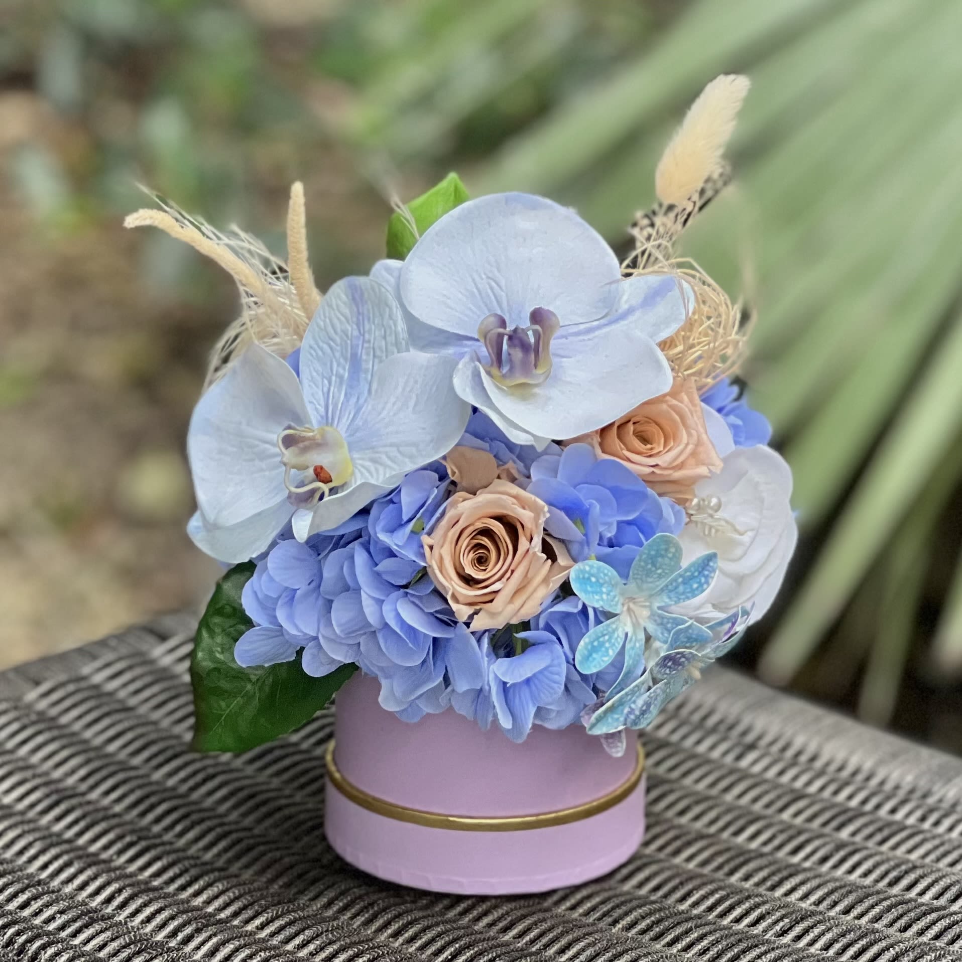 Mini Blue Orchid Bloom Mini Blue Orchid Bloom - Experience the tranquility of everlasting elegance with Blue Orchid Bloom. This mini arrangement brings together the soft, cool tones of blue hydrangea, the warm subtlety of beige roses, and the pristine beauty of white orchids. Dried elements delicately accent the design, adding texture and depth to this charming, compact arrangement. Presented in a petite, stylish box, Eternal Serenity is the perfect way to send a touch of calm and beauty. With its preserved flowers, this eco-friendly, low-maintenance gift offers lasting charm — a timeless keepsake for any occasion.