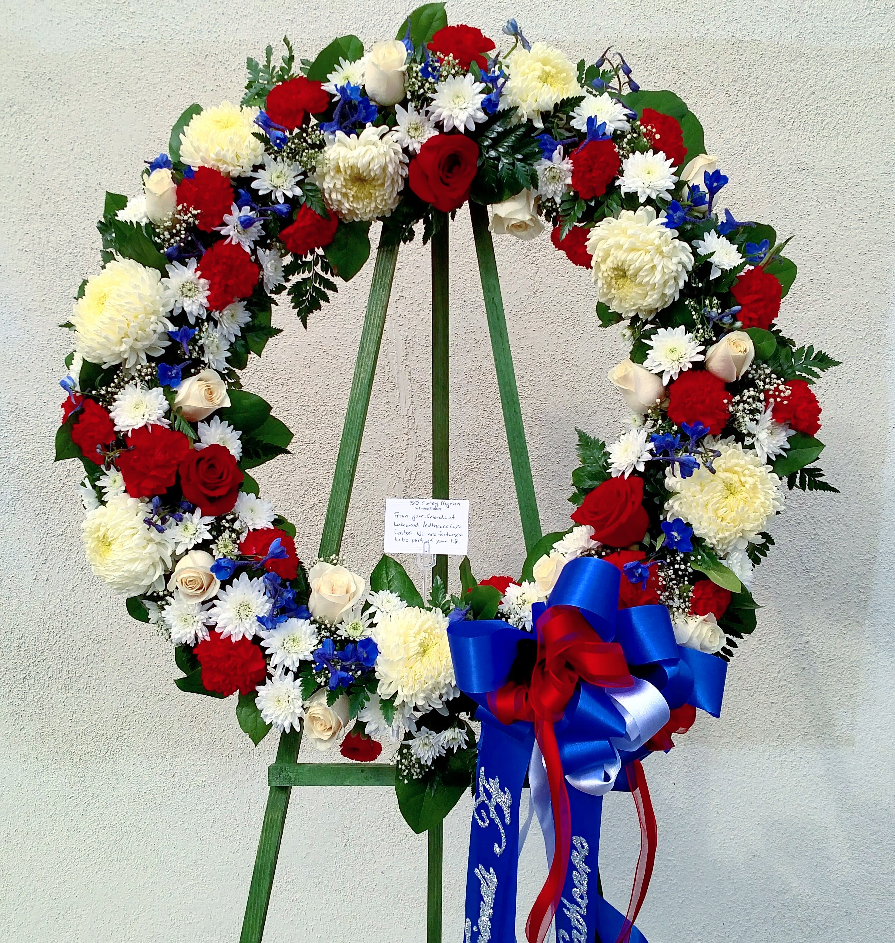 Tribute Wreath - Vibrant veterans red, white and blue wreath china mums, roses, carnations, and blue delphinium.  A big beautiful bow with hanging tails included. To add wording please add by searching &quot;Sympathy Banner&quot;.