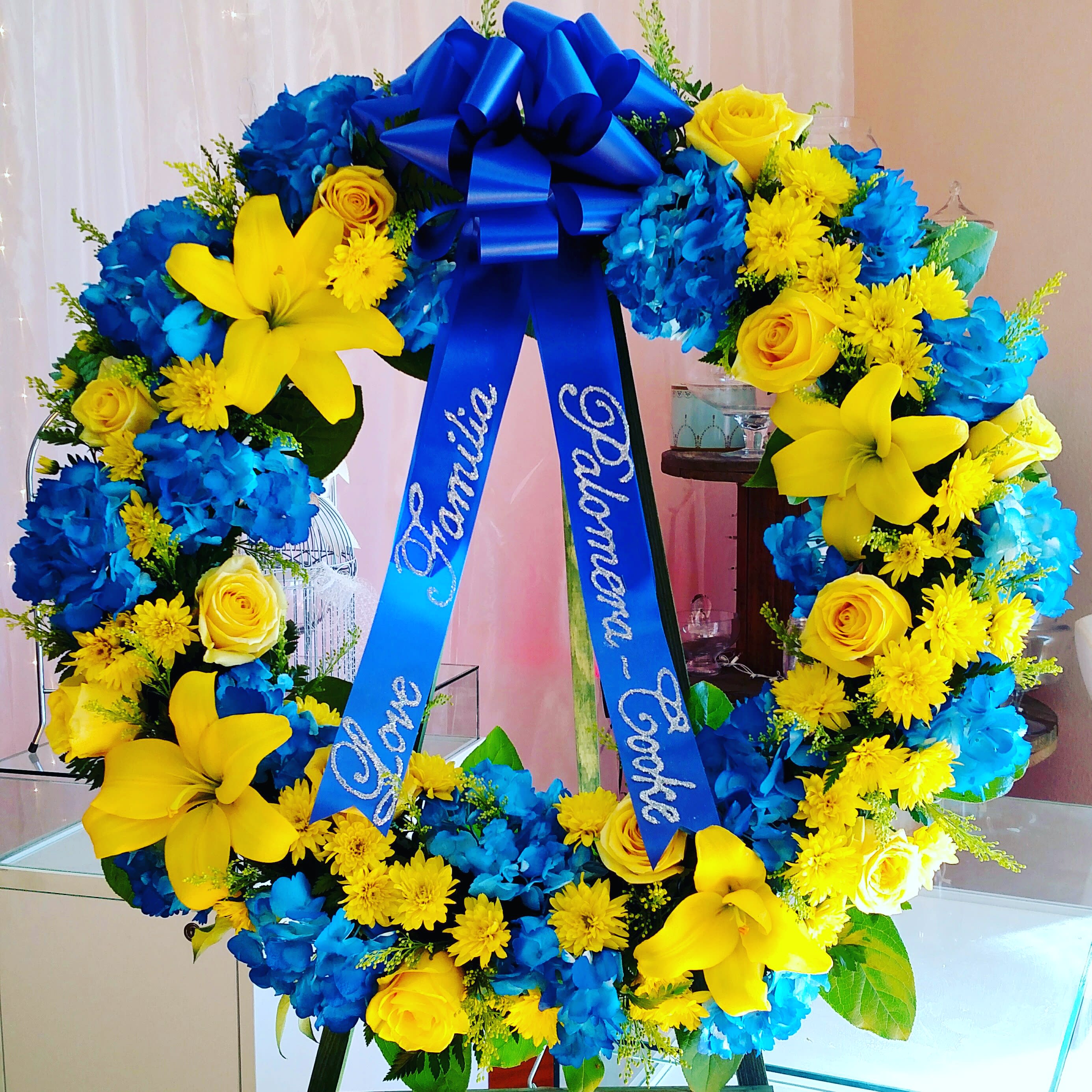 Yellow Lily And Rose Wreath - Bright yellow lilies and roses with deep blue hydrangea and yellow accent flowers that will bring brightness to the services. For a custom banner, please add by searching &quot;Sympathy Banner&quot;.