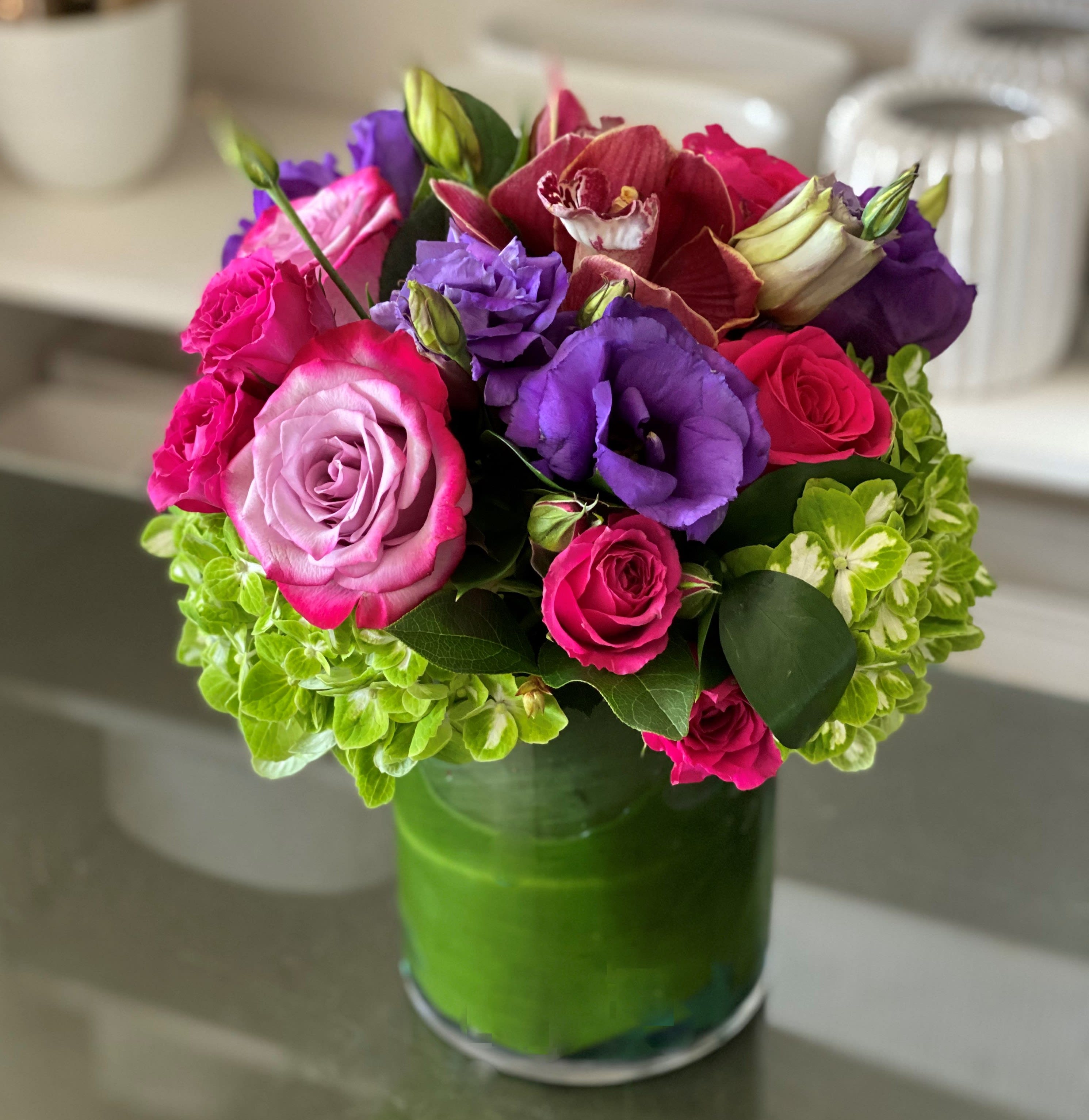 Olvera  - The Olvera Arrangement is filled in Bold Bright Color florals that you can  send in any occasion all year round. Lavender Roses, Purple Lisianthus, Green Hydrangeas and burgundy Cymbidium Orchids in a 6x5 clear cylinder vase. 