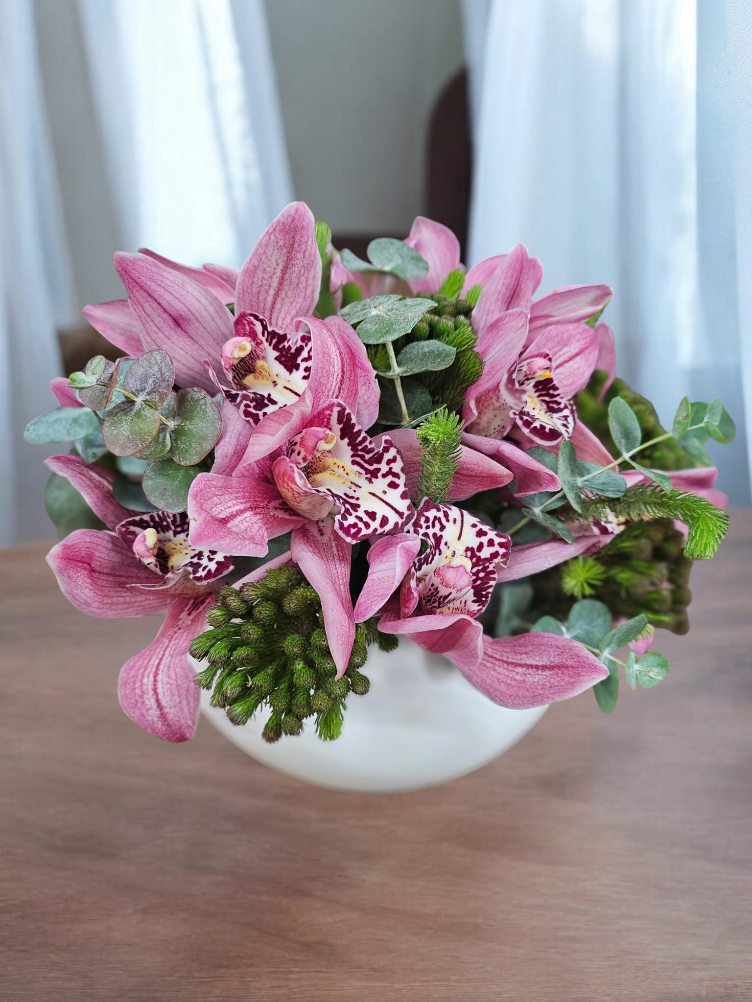Luxurious Pink Orchid Harmony in a Modern White Vase - This elegant arrangement features exquisite pink cymbidium orchids complemented by lush greenery and delicate eucalyptus accents. Artfully arranged in a sleek white ceramic vase, this design radiates sophistication and charm, making it a perfect centerpiece or gift for any occasion.  Premium Flowers: Beautiful cymbidium orchids and seasonal greenery.  Presentation: Displayed in a chic white round vase for a contemporary look.  Occasions: Ideal for anniversaries, birthdays, housewarmings, or as a thoughtful gesture to brighten someone's day  Each arrangement is meticulously handcrafted to ensure a stunning display of beauty and freshness. Add a touch of luxury and elegance to any space with this timeless floral piece.