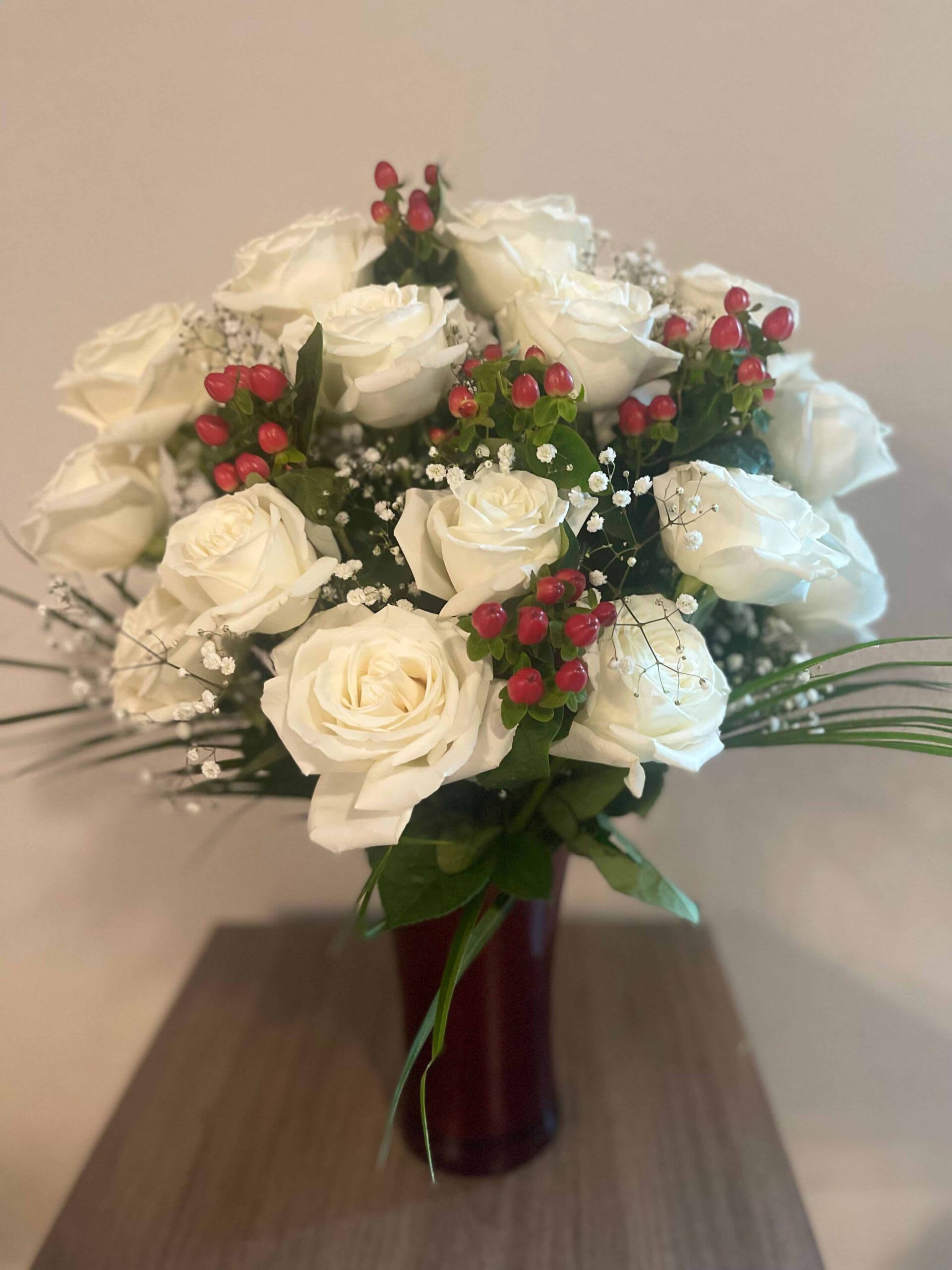 White Christmas - Celebrate the most joyous holiday with this enchanting white bouquet. The purity of the Christmas season is reflected in these delicate twenty-four long-stem white roses. Red hypericum, Baby’s Breath, and assorted greenery are integrated to add depth. The flowers are artfully exhibited in a tall red festive vase.