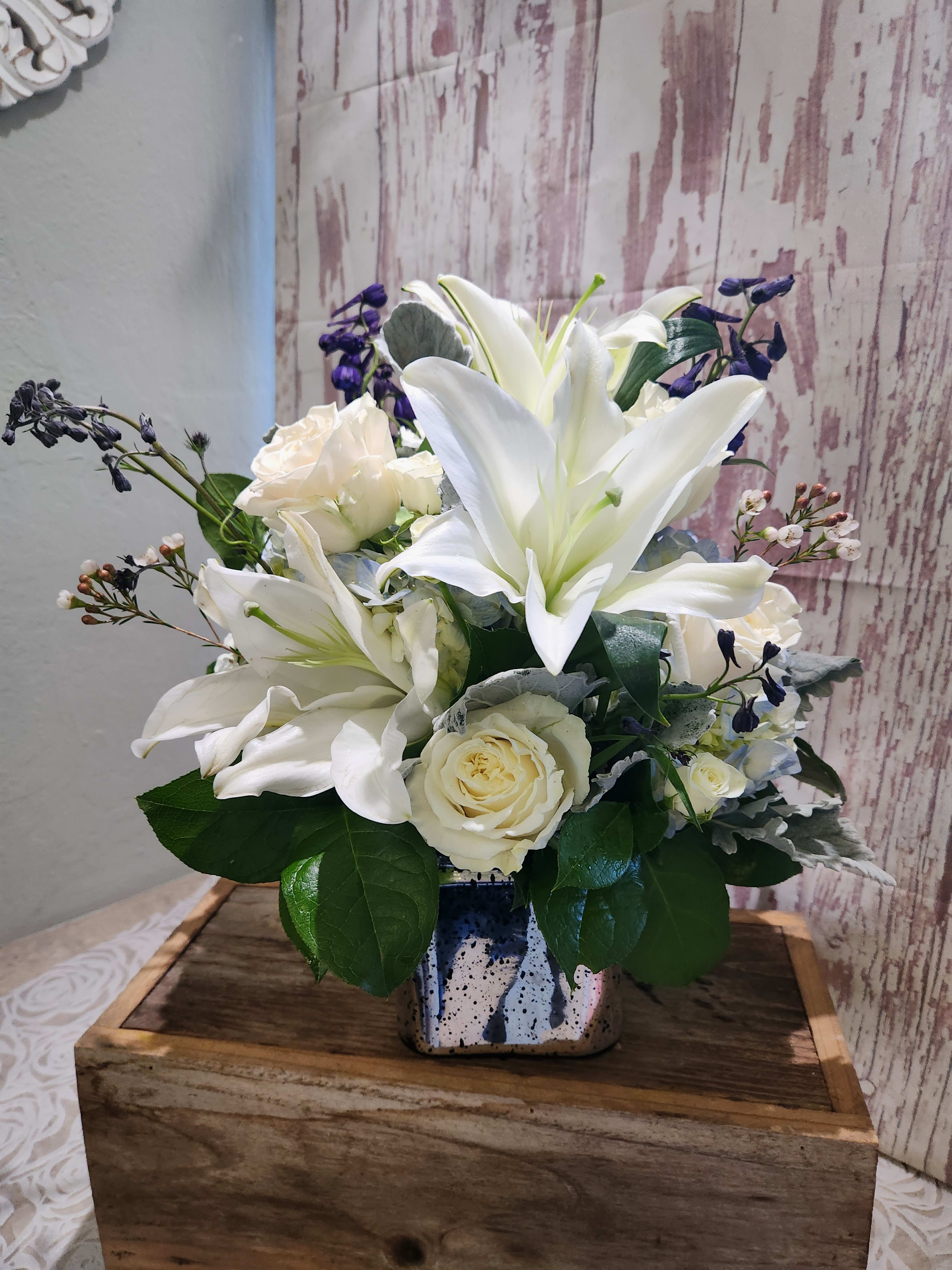 Seaside Centerpiece -  A beautiful mixed blue and white hydrangeas, delphinium, white creamy roses, white lilies. Will be delivered a cube or cylinder vase.  (VASE  MAY VARY due to availability) it's an elegant choice for weddings and showers. Standard will be one side Deluxe size is all around  Pictured is a Deluxe Size  