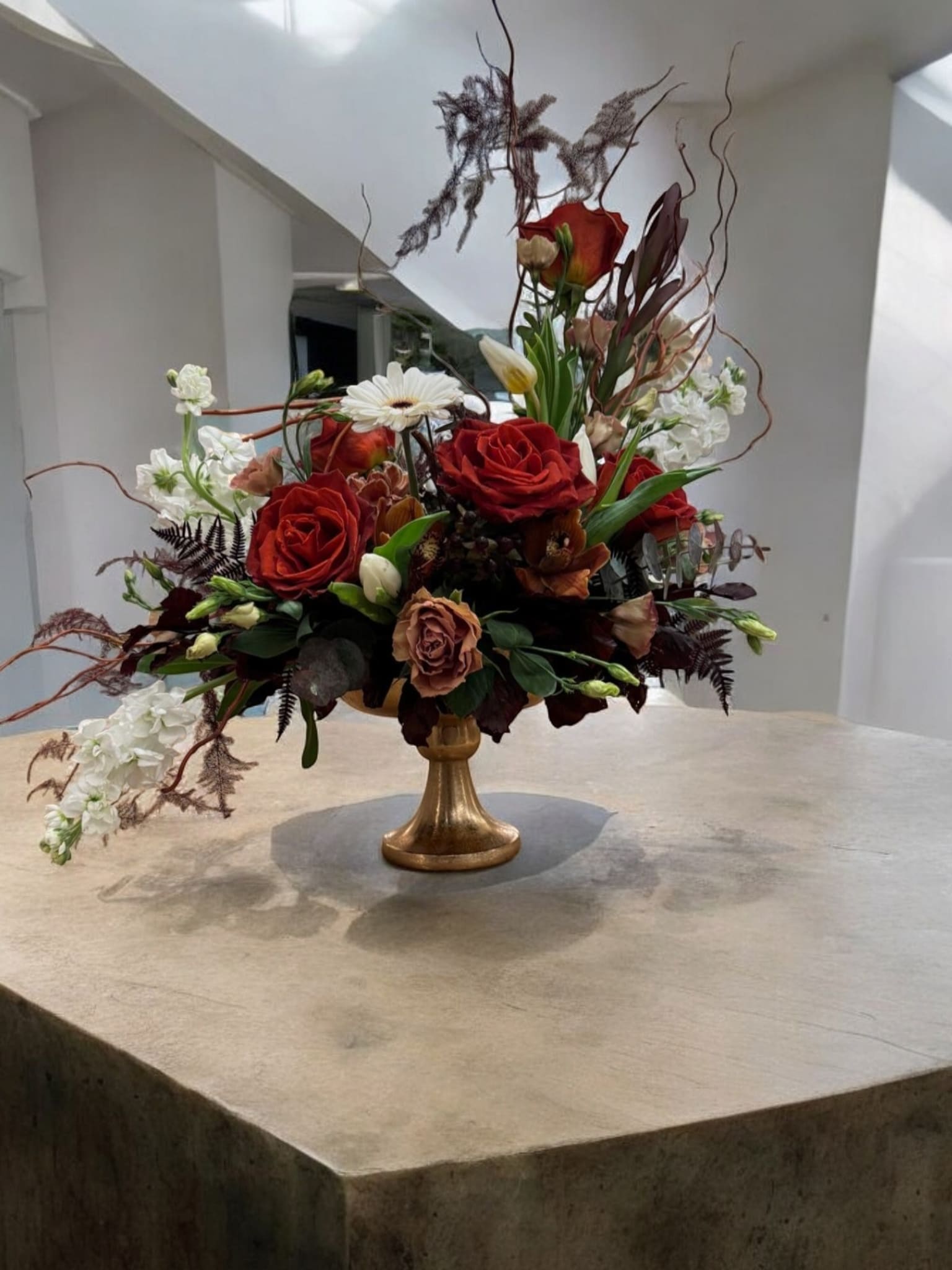 Elegant Autumn Harvest Floral Arrangement - This luxurious centerpiece features a striking combination of deep red roses, creamy white blooms, and earthy accents, arranged in a sophisticated golden pedestal vase. Designed with artistic flair, the arrangement includes unique textural elements like curly willow branches and seasonal foliage, adding height and drama to the composition. Perfect for upscale events, intimate dinners, or as a show-stopping gift, this design captures the essence of timeless elegance and autumnal warmth.  Premium Flowers: Red roses, white snapdragons, gerbera daisies, and seasonal accent blooms.  Vase Style: A classic golden pedestal vase for added grandeur.  Occasions: Ideal for weddings, anniversaries, formal gatherings, or as a luxurious gift for someone special.  Size: Approximately 18–22 inches tall and wide, making it a stunning focal point.