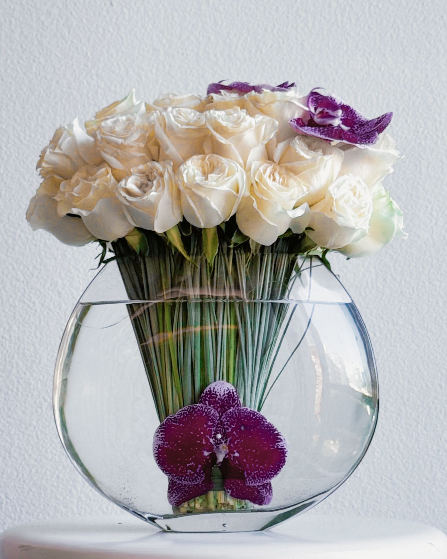 Orchid Elegance - This bouquet is the epitome of timeless elegance and deep emotion. The pure ivory roses symbolize everlasting love and devotion, while the striking purple orchids add a touch of exotic passion. Arranged in a sleek glass vase, it’s a harmonious blend of classic beauty and modern sophistication, making it the perfect gift to express feelings that words cannot capture.  Occasions for Gifting: Anniversaries: Celebrate a love that grows stronger with time. Weddings: A meaningful centerpiece or gift for the happy couple. Milestones: Mark a significant life event, such as a promotion or graduation. Romantic gestures: A declaration of love or a thoughtful surprise for someone special. Condolences: A symbol of peace, comfort, and eternal beauty. 