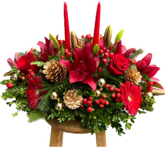 Red festivities  - This stunning holiday arrangement is a vibrant celebration of the season, featuring a beautiful mix of red flowers—lush lilies, classic roses, and cheerful gerbera daisies. Their deep, rich hues are perfectly complemented by the natural charm of pine cones, adding an earthy, rustic touch. Golden ornaments glimmer amidst the blooms, bringing a festive, elegant sparkle to the design. Atop the arrangement, a pair of flickering candles offer a warm, inviting glow, creating a cozy, holiday atmosphere. This striking centerpiece effortlessly combines the beauty of nature with the magic of the season, making it an ideal addition to any holiday table or festive gathering.