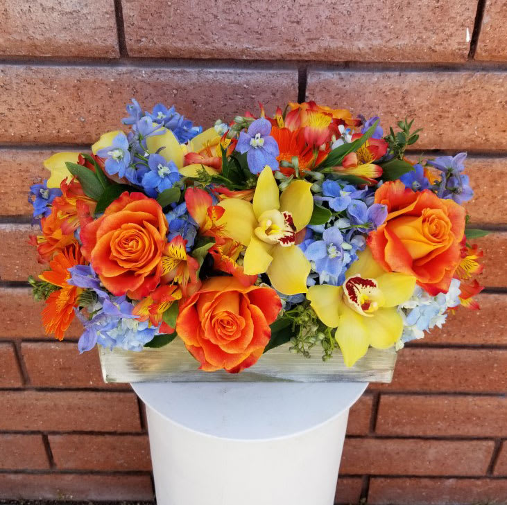 Florescence - This lovely, colorful, fresh, and unique arrangement features hydrangeas, delphinium, premium roses, gerbera daisies, cymbidium orchid and alstroemerias, all hand arranged in a wood box. Overall size of the arrangement is approximately 16in (W) x 9in (H).  Arrangement Features: 6 Orange Roses, 4 Yellow Cymbidium Orchid Blooms, 5 Delphinium, 5 Orange Alstroemerias, 3 Orange Gerbera Daisies, 3 Blue Hydrangeas  Care Tips: This centerpiece is anchored in a floral foam and can stay vivid and beautiful for days with the proper care. OASIS floral foam retains water and keeps the stems of the flowers hydrated. The key is to make sure the foam stays moist, so add water accordingly. Keep your floral arrangement in a location without direct sunlight and away from extreme heat. For best results do not place the centerpiece near a drafty door or near a source of heat.