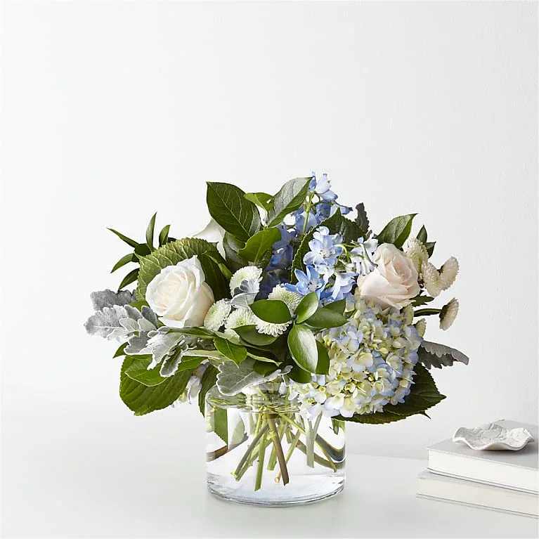 Light Blue Skies Bouquet - Let this uplifting arrangement be reminders of the clear skies ahead. Capturing the feeling of hope and renewal that a new day brings, this bouquet is composed of voluminous hydrangea blooms, vibrant belladonna delphinium and white pompon buttons to refresh anyone's mood.