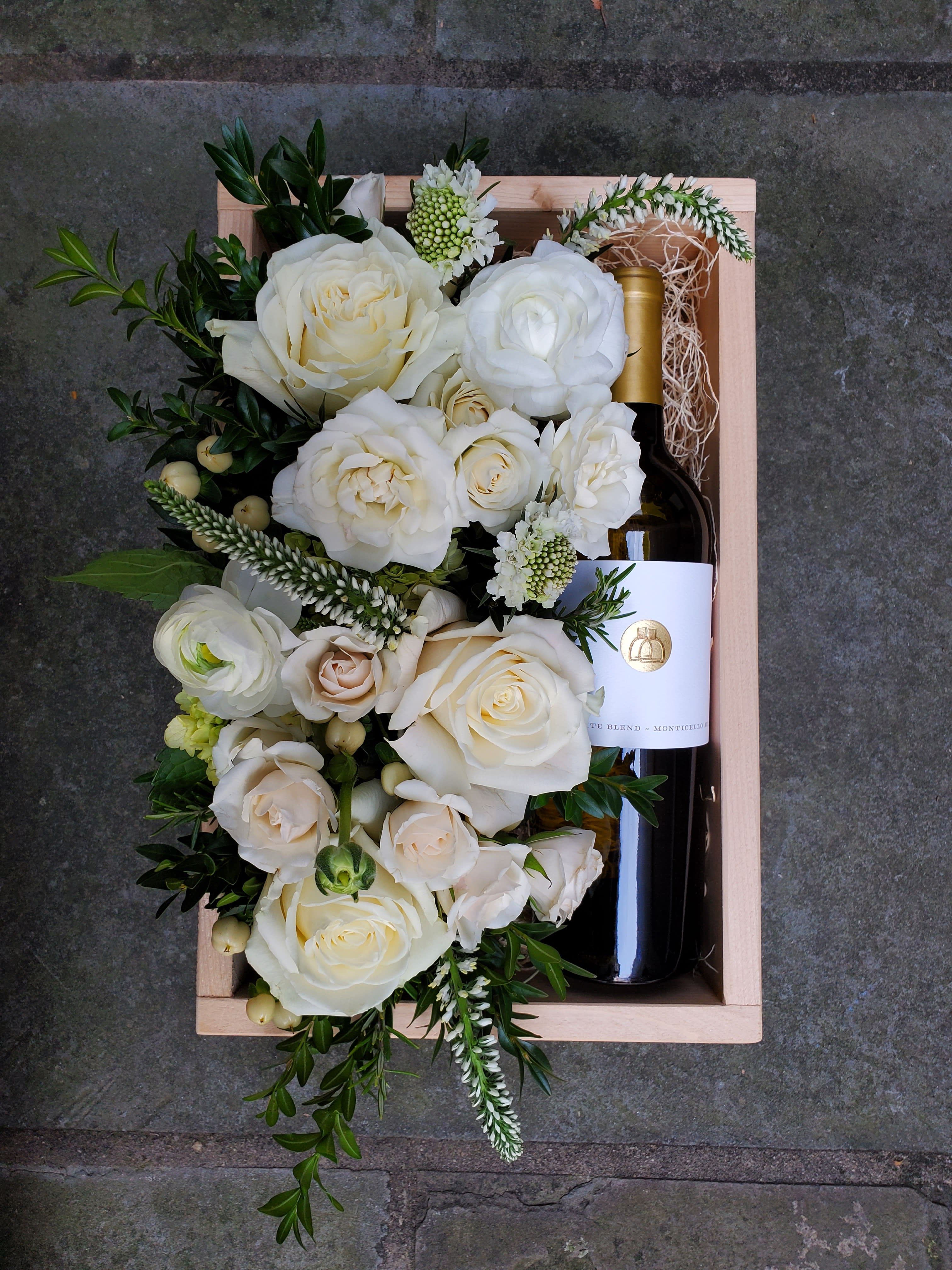 White Wine Blend Gift Box White Wine Blend Gift Box - A beautiful wooden gift box, locally hand-crafted from reclaimed wood. A bottle of Southwest Mountains Vineyards' White Blend wine rests on a bed of Spanish moss, alongside a beautiful bed of flowers in the pavé style. You may specify a color for this arrangement if desired. Wine Description: White Blend from Southwest Mountains Vineyards is a well-balanced wine made from a blend of local favorites of the Keswick-based winery, Viognier and Petit Manseng. With notes of peach, apricot, and orange blossom, this medium-bodied yet lively wine finishes relatively soft on the palate. Before placing your order, please ensure that the recipient is above 21 years of age and will be home to receive the delivery, as ABC regulation requires that we check photo ID upon delivery. If no one is present at the delivery site, delivery cannot be completed. Box measures 8.5x11.5".