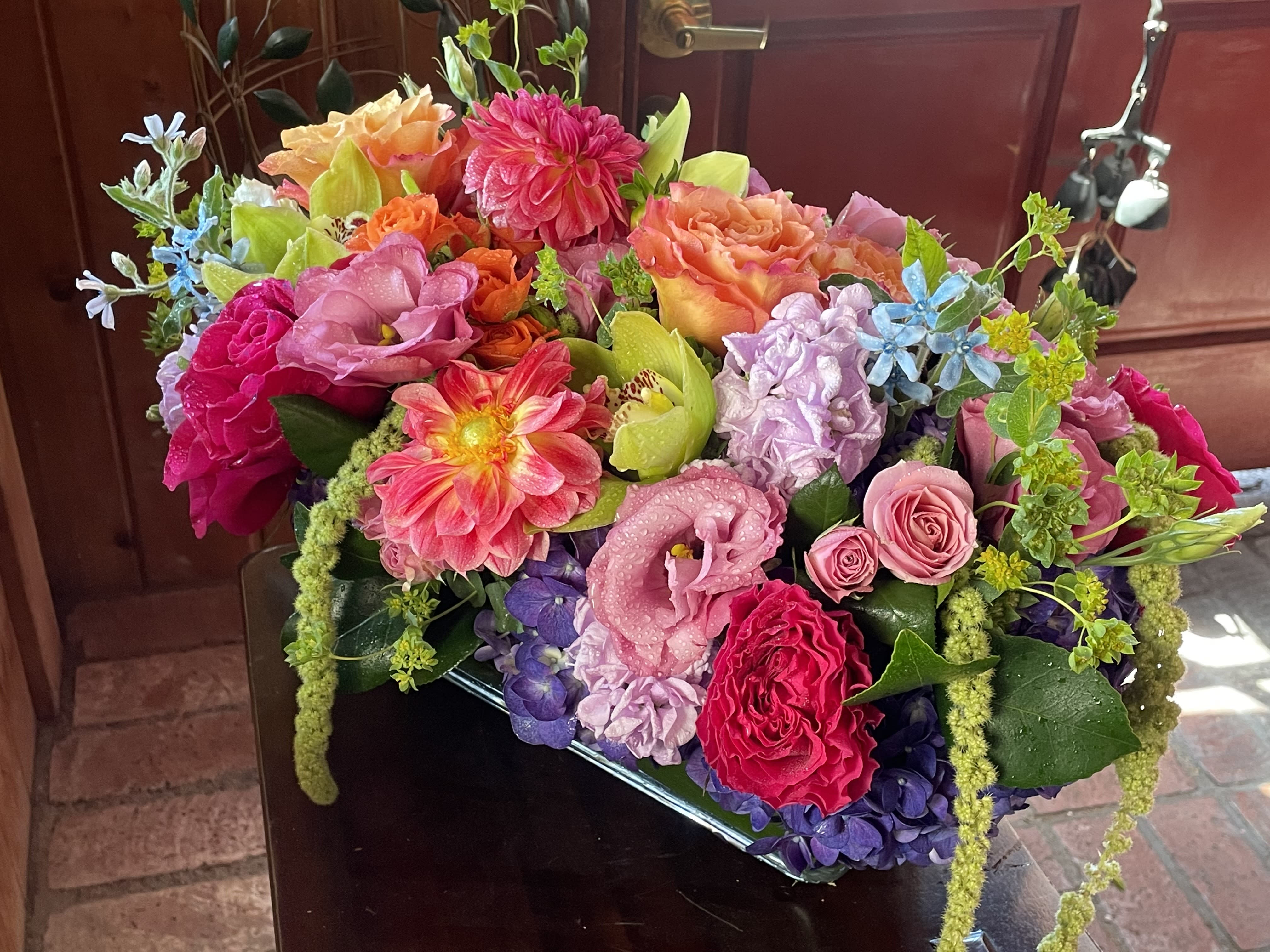 Midsummer Roll -  This stunning, long and low vase arrangement of seasonal dahlias is here to steal the show at your next gathering. With its vibrant colors and perfectly balanced design, it's sure to be the centerpiece that has everyone saying, &quot;Wow!&quot; Whether you're hosting a summer soiree or just want to add some pizzazz to your dining table, the Midsummer Roll is the floral superstar you've been waiting for. Grab one now before they roll away!  Approximately 20&quot; long by 8&quot; wide in a long and low rectangle clear vase.     Features dahlias, hanging amaranthus, orchids, seasonal greenery, blue tweedia, roses, and purple local hydrangea. 