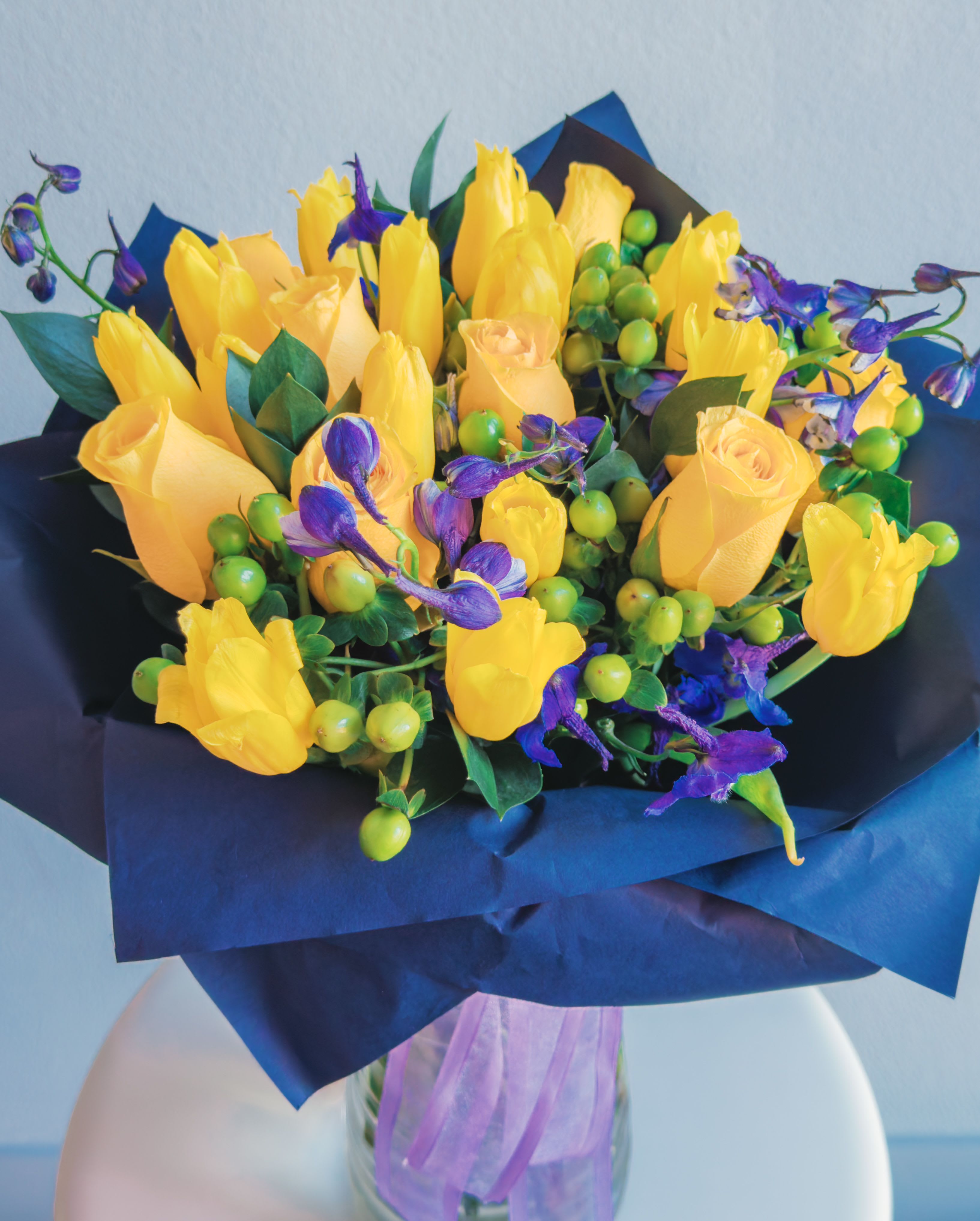 Sunny Soul - This bouquet is a celebration of contrasts and harmony! Vibrant yellow roses and tulips radiate energy, while the deep purple delphiniums create a striking color contrast, bringing depth and elegance. Green hypericum berries add playful texture, and the lush greenery ties everything together in a modern, bold arrangement. Wrapped in navy blue paper with a soft purple ribbon, this bouquet feels like a ray of sunshine captured in a sophisticated frame. 
