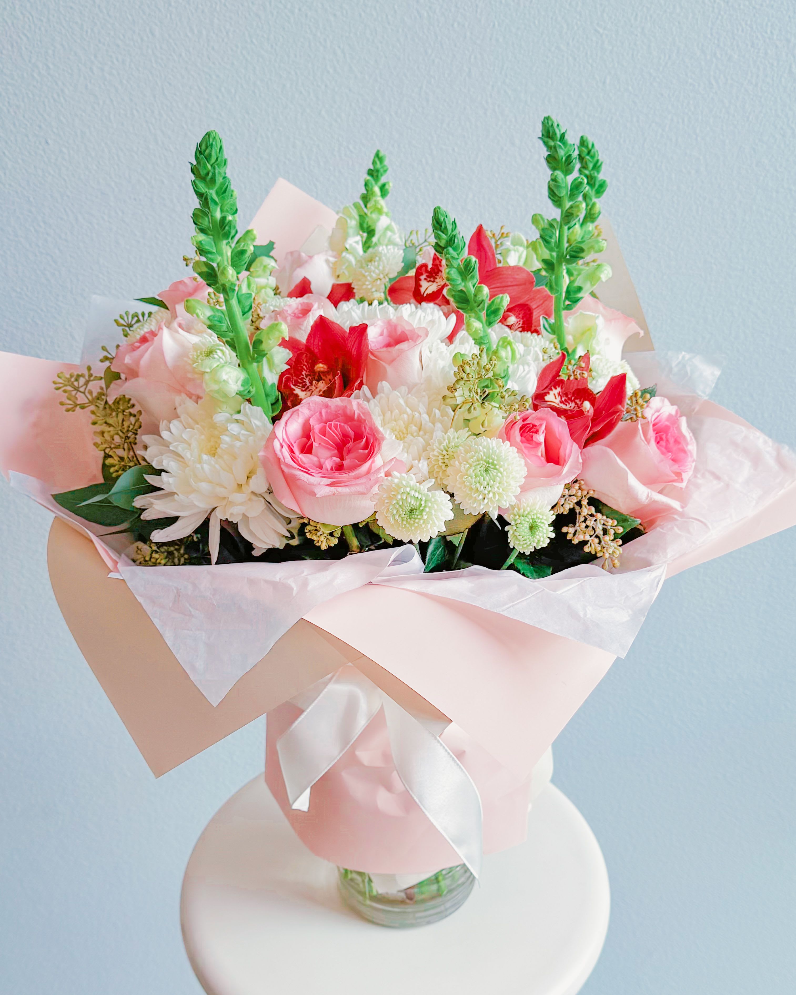 Flirty Fleurs - Alright, this bouquet is what I’d call a total showstopper — the perfect mix of soft pink romance and pops of bold red to keep it spicy. We’ve got roses that are blushing like a first date, delicate orchids that bring drama, and snapdragons adding that playful height. It’s wrapped up in dreamy pastel packaging, giving it that modern, fresh-out-of-NYC look. You could give this beauty to celebrate love, friendship, or just to say, “Hey, you’re amazing!”