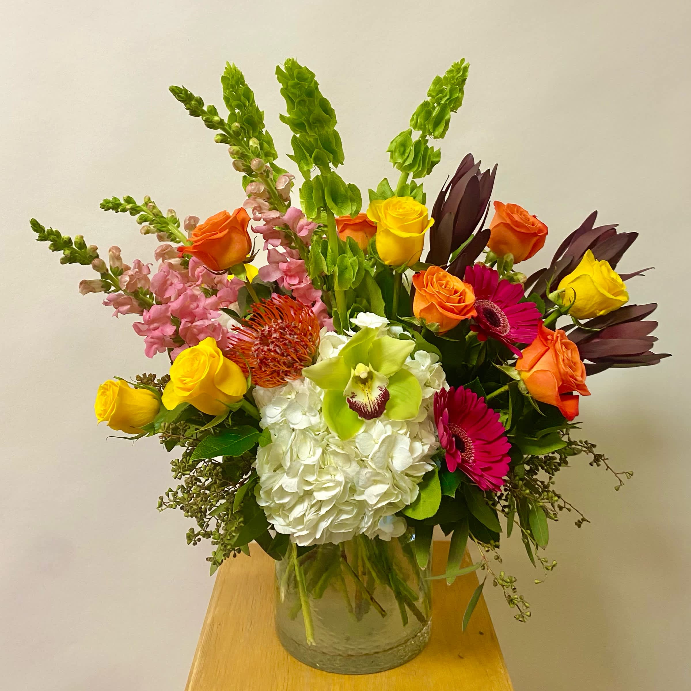 Charmed Charmed - This Medium Tall Lush Arrangement includes White Hydrangeas, Pincushions, Green or Pink Cymbidium Orchid Blooms, Bells of Ireland, Light Pink or Orange Snapdragons, Yellow Roses, Orange Roses, Hop Pink Gerbera Daisies, Safari Sunsets in a Premium Glass Vase.