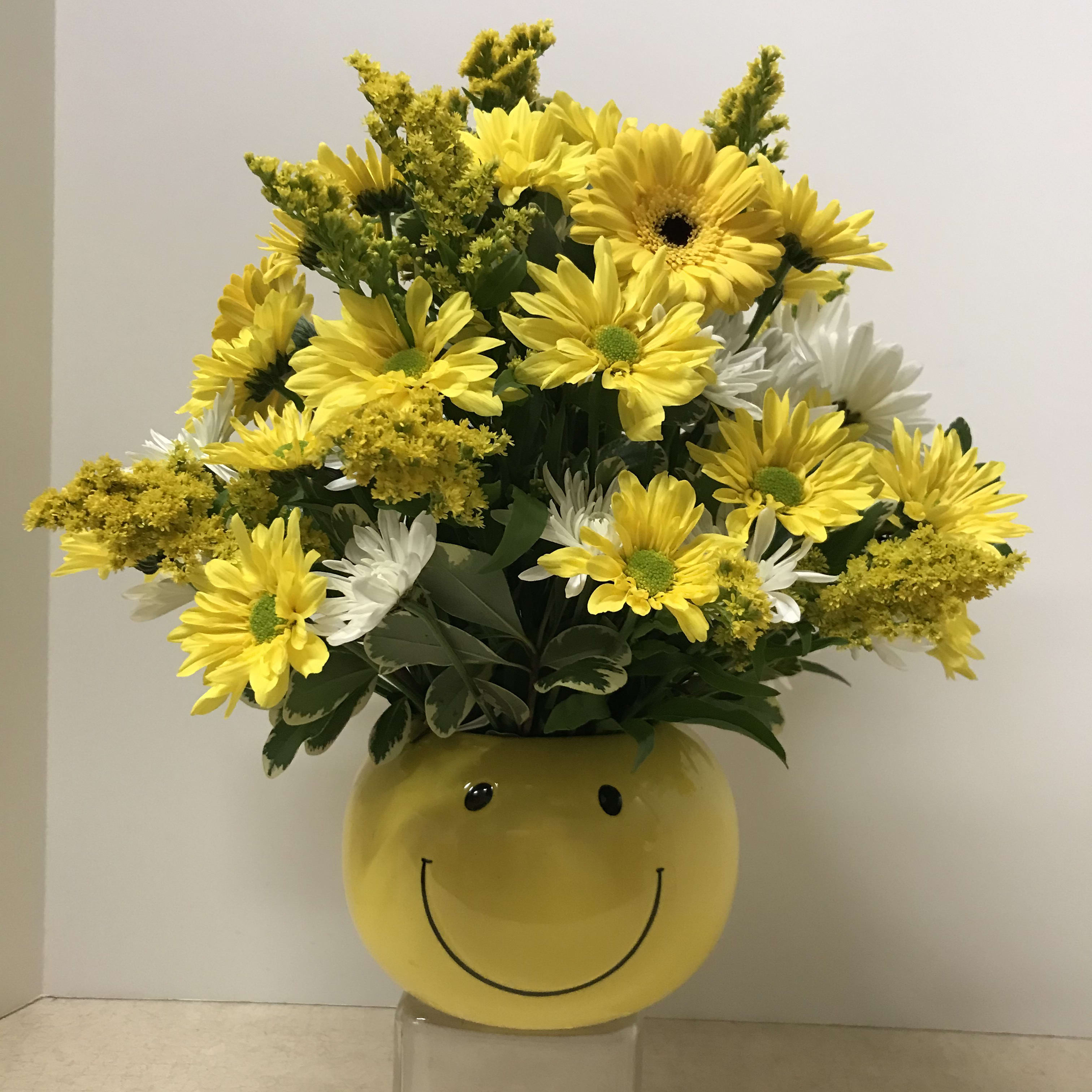 "A Smile and Blossom Bouquet" "A Smile and Blossom Bouquet" - This bright and cheerful beautiful bouquet features a ceramic yellow bowl adorned with a happy smiley face and filled with a variety of seasonal flowers. Yellow daisies, white cushion pompon mums, solidago, gerbera daisies, and assorted greens complete this happy centerpiece that is sure to brighten up any day!