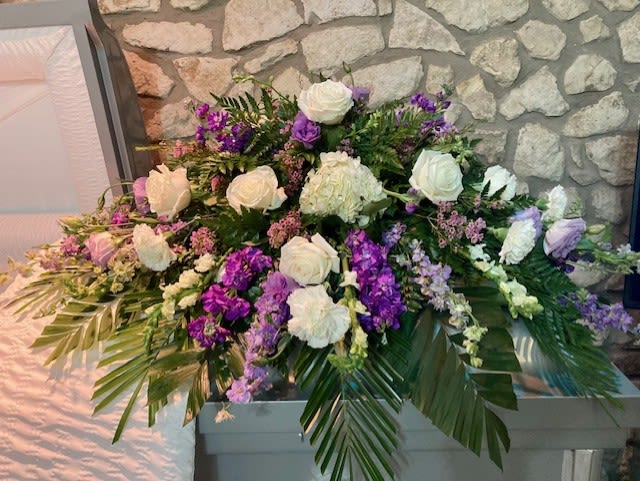 Purple and White Casket Blanket - This is a beautiful purple and white casket piece made with a variation of flowers. Please note that we will try our best to make it using the same colors/style/flowers however some flowers are only available in a season Also, please note that often due to the nature of funerals happening without a lot of time to plan, we are only able to reserve the floral that is available to our wholesalers during that timeframe. If a funeral is planned for the end of week, floral may be limited. It is best to order a casket spray over the phone or in person. 