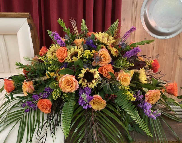 Sunflower and Free Spirit Casket Spray - Sunflowers mixed with Fall favorites make this perfect for either a man or a woman. Floral availability on casket sprays vary depending on season and the amount of notice given for floral availability. We will do our best to keep the overall style in place when ordering. This will be sized for both an open or a closed casket.  Please note that we will try our best to make it using the same colors/style/flowers however some flowers are only available in a season Also, please note that often due to the nature of funerals happening without a lot of time to plan, we are only able to reserve the floral that is available to our wholesalers during that timeframe. If a funeral is planned for the end of week, floral may be limited. It is best to order a casket spray over the phone or in person. 