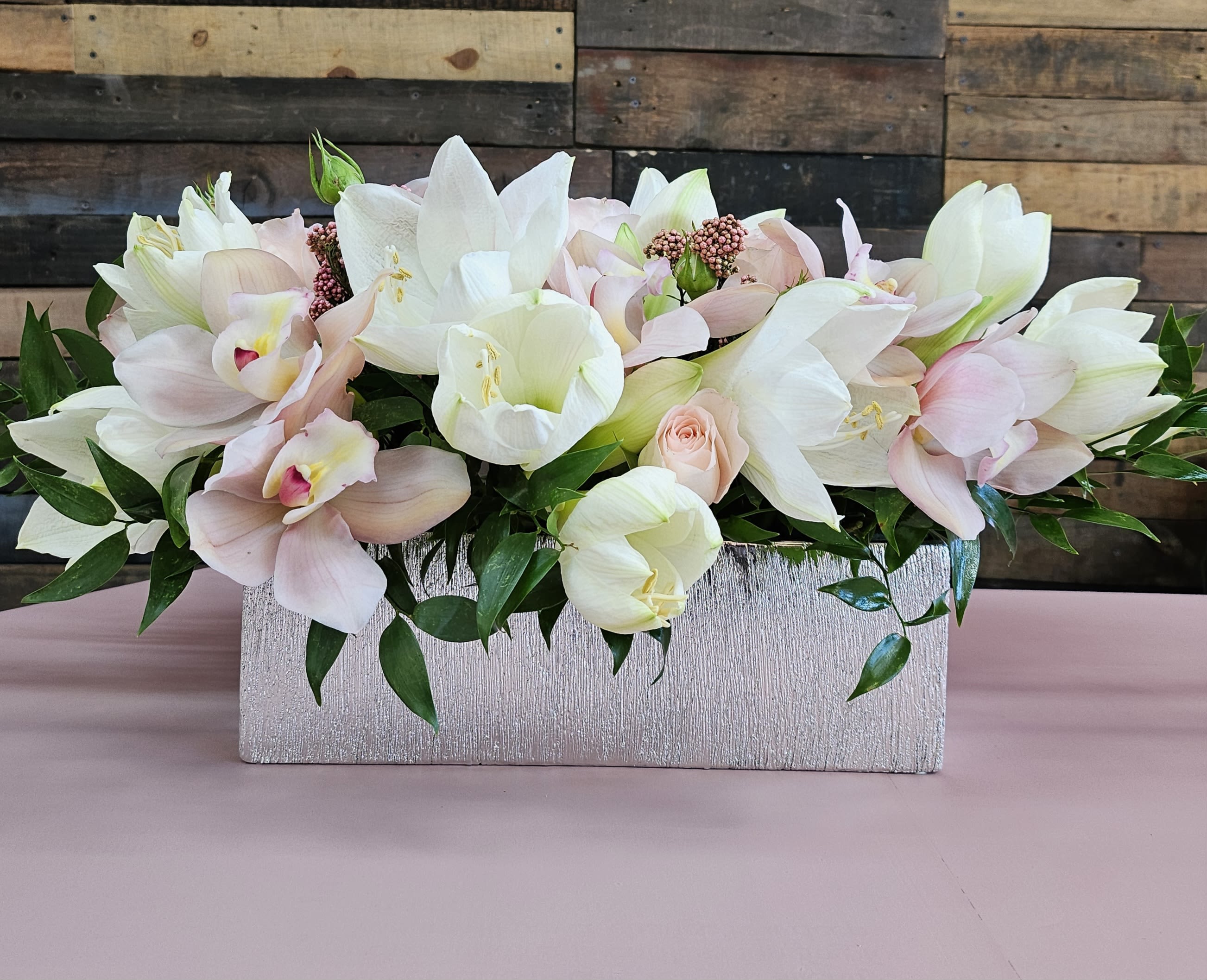 Soft Blush Radiance Floral Arrangement - Elevate any setting with this exquisite arrangement featuring a harmonious blend of soft blush tones and elegant whites. Showcasing delicate amaryllis, pink cymbidium orchids, blush roses, and lush greenery, this arrangement is designed in a textured silver vase for a modern yet timeless look. Perfect for creating a sophisticated ambiance for any occasion.  Premium Flowers: Amaryllis, cymbidium orchids, blush roses, and seasonal accents.  Vase Style: A chic textured silver rectangular vase that adds a touch of luxury.  Occasions: Ideal for weddings, anniversaries, celebrations, or as a centerpiece for elegant gatherings.  Size: Approximately 20–24 inches in length and 10–12 inches tall, offering a grand and impactful presence.  Care Instructions: Keep in a cool location, refresh water every 2–3 days, and avoid direct sunlight to maintain freshness.   This arrangement is the perfect blend of beauty and sophistication, making it a standout addition to any décor or event