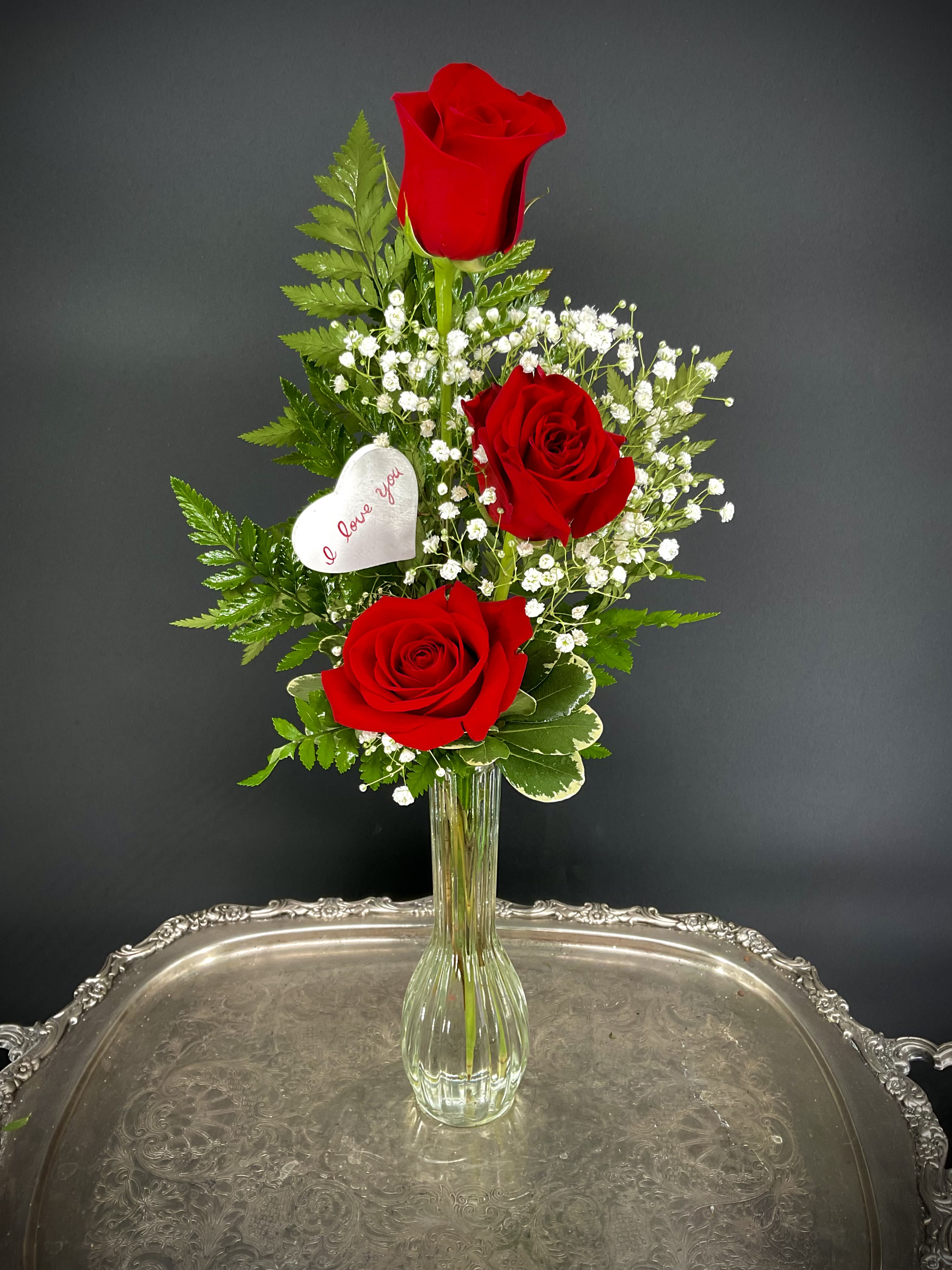 Three Red Roses in a Bud Vase - Send a cute and sweet red rose bud vase to your love, your friend, your secret crush or to your relative for Valentine's Day.  Three red roses are accented with greenery, baby's breath and an &quot;I Love You&quot; heart shaped wooden Valentine's Day keepsake.  Sending this arrangement is a lovely gesture and shows that you are thinking of them. 
