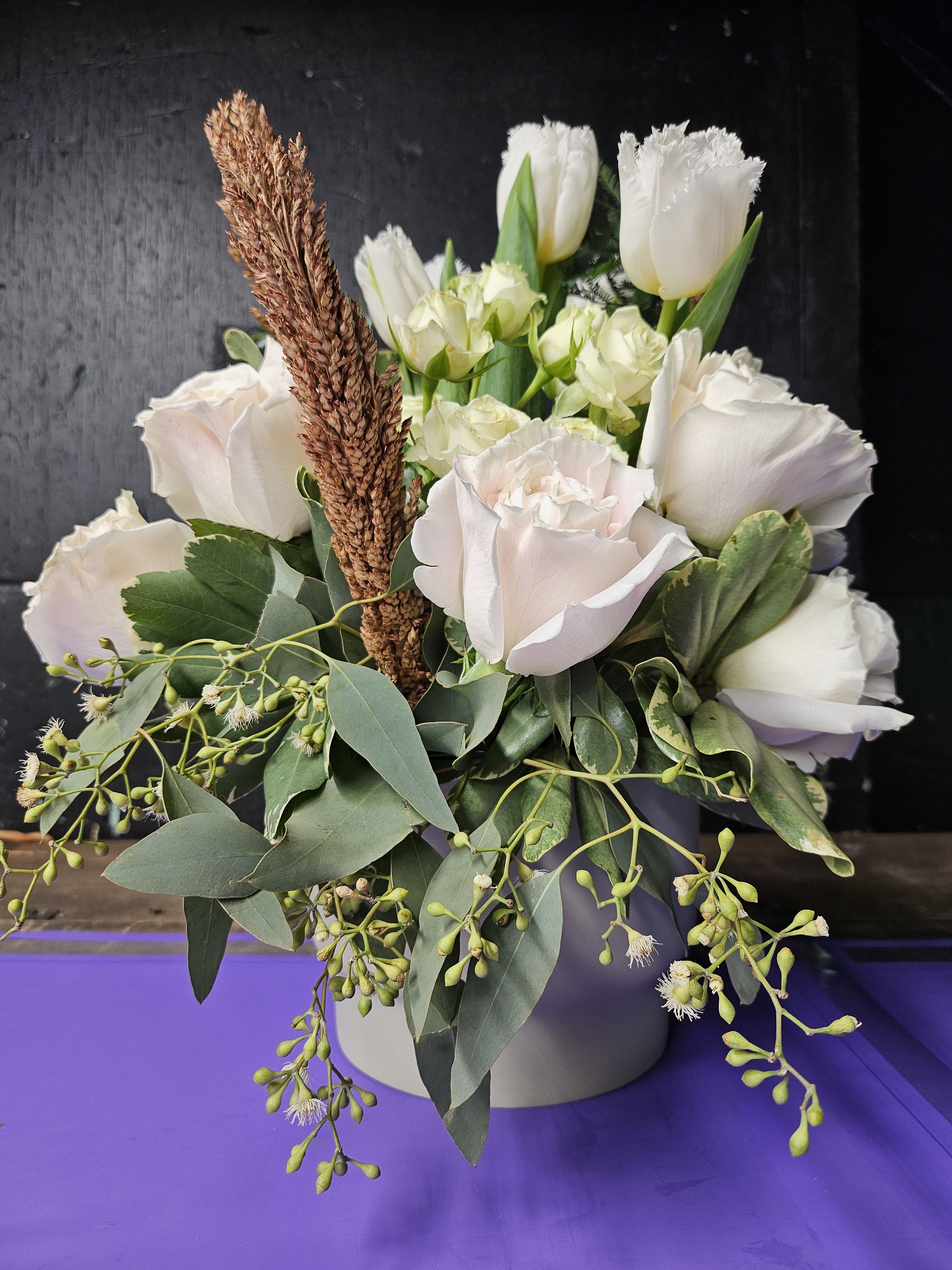 White Elegance: Roses, Tulips &amp; Eucalyptus in Modern Vase - Embrace the serene beauty of this exquisite arrangement, featuring creamy white Roses, delicate fringed Tulips, and elegant Spray Roses. Accentuated with lush Eucalyptus greenery and dried textured wheat for a rustic yet sophisticated touch, this bouquet is arranged in a modern, neutral-toned vase, making it perfect for any setting. Whether for a celebration, a heartfelt gift, or a peaceful addition to your home, this arrangement exudes timeless grace and natural charm.