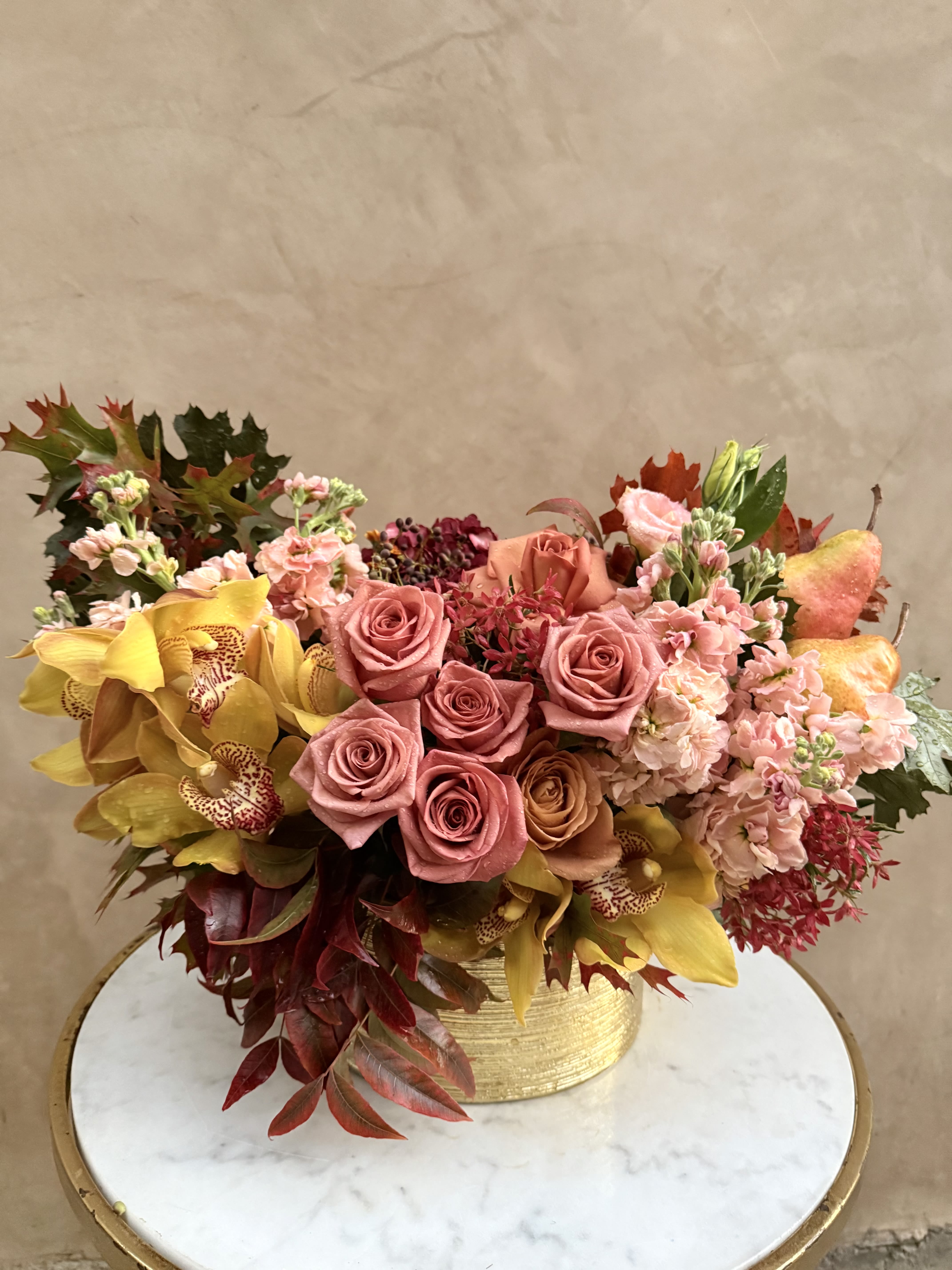 Thanksgiving Harvest  - Celebrate the beauty of fall with this stunning arrangement featuring a warm palette of soft pink and golden hues. Arrangement may include but not subject to; velvety peach roses, golden cymbidium orchids, and delicate blush stock flowers, accented with rich red foliage and seasonal textures. Complemented by crisp apples and arranged in a chic. This design radiates rustic charm and elegance. Perfect for bringing the warmth of autumn indoors, it makes an ideal centerpiece for Thanksgiving gatherings, fall weddings, or seasonal décor.