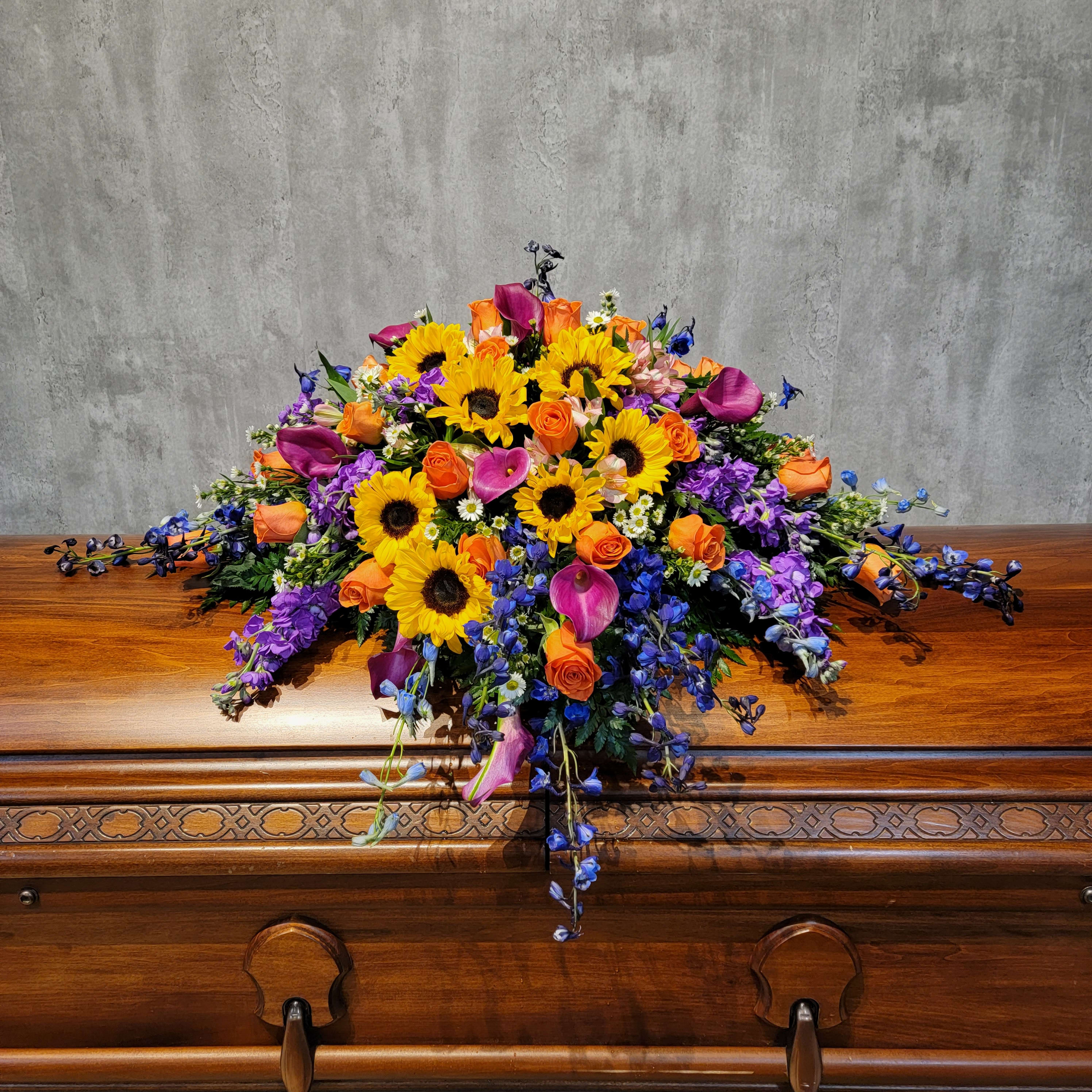 Country Meadows Casket Spray - Gorgeous combination of calla lilies, roses, delphinium and sunflowers make for this beautiful country meadows casket spray. This casket spray will fit on the half casket when open, to cover the full casket when closed please choose the deluxe.