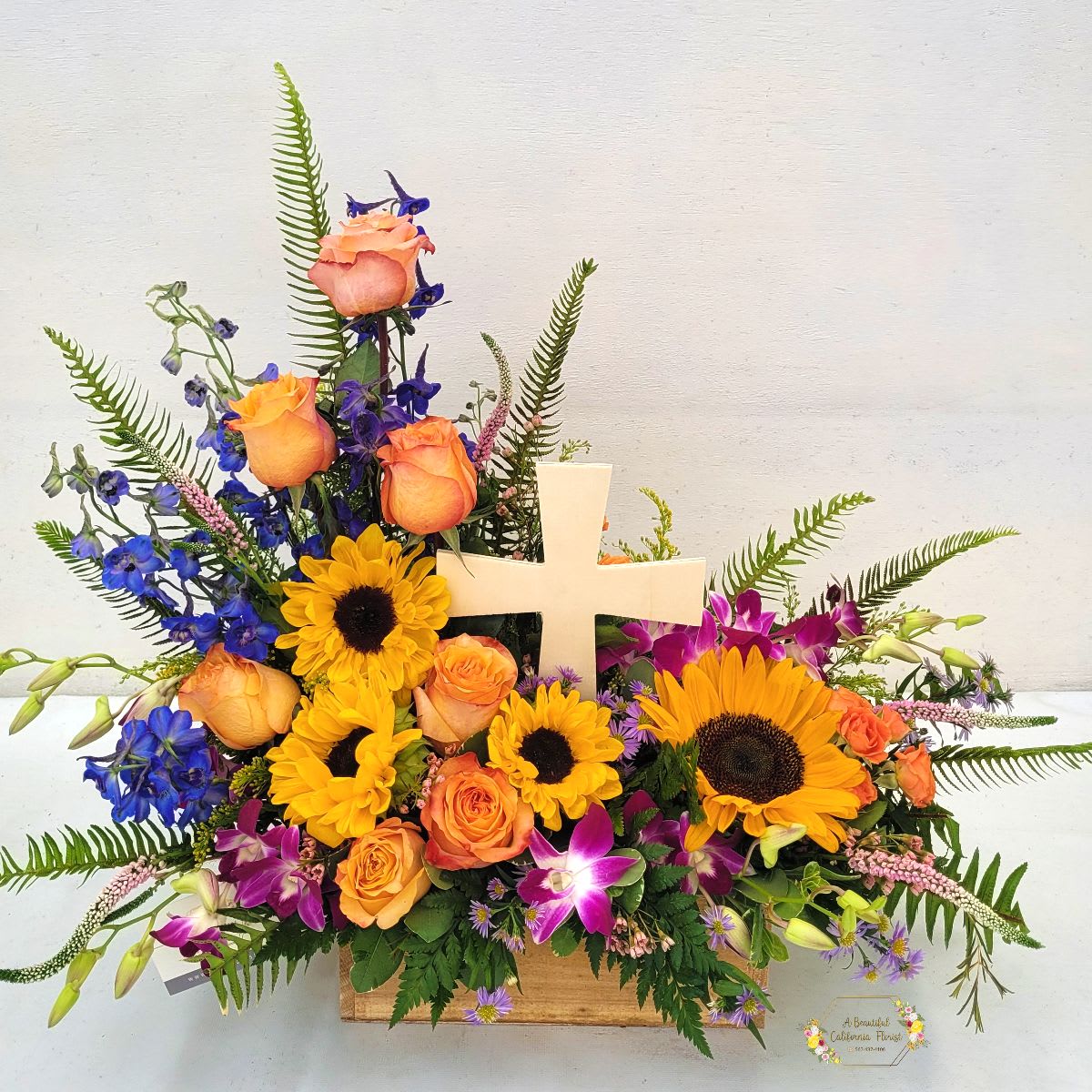 Eternal Hope Eternal Hope - Eternal Hope A timeless tribute of solace and remembrance, Eternal Hope is a breathtaking arrangement that embodies the enduring spirit of hope. Radiant sunflowers symbolize warmth and optimism, while elegant roses convey deep sympathy and love. The serene blue delphinium and exotic bombay orchids add a touch of tranquility and grace. Lush greens frame the centerpiece, a wooden cross that serves as a poignant reminder of faith and eternal life. This exquisite arrangement is presented in a rectangular wooden container, offering a lasting keepsake of comfort and support. Approximate size 18” T x 22” W