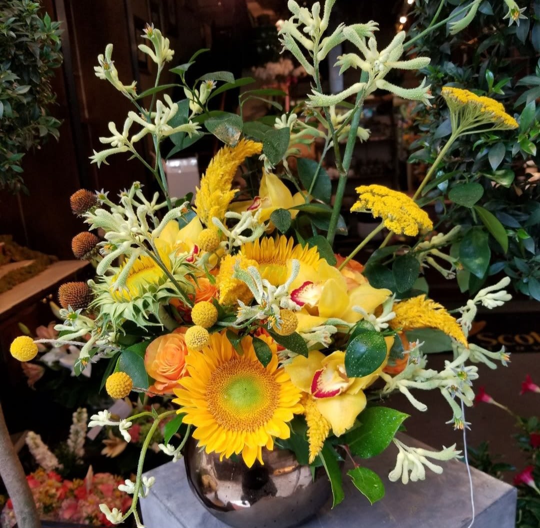 Wild Sunflower Medley - The Wild Sunflower Medley arrangement is a vibrant celebration of nature’s golden hues, perfect for capturing the wild beauty of autumn. Featuring radiant sunflowers paired with delicate yellow orchids, this bouquet is accented with seasonal fillers that add texture and depth. The arrangement is completed with fall foliage in rich tones of amber, russet, and gold, evoking the crisp beauty of an autumn day. With its untamed charm and sunlit warmth, the Wild Sunflower Medley is ideal for bringing the spirit of the season into your home or sharing a touch of autumn’s magic with someone special.