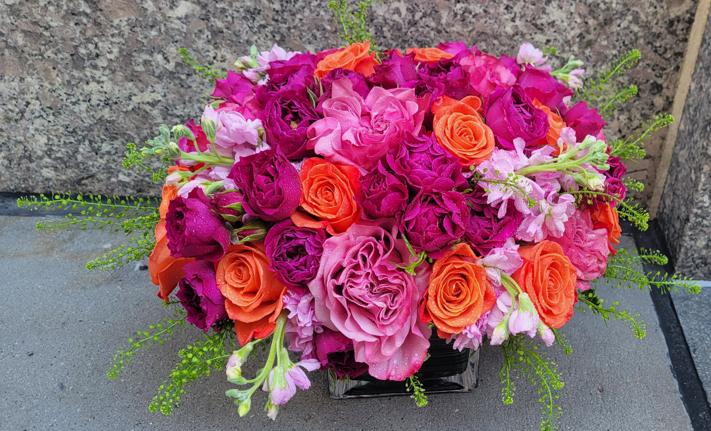 Ruby Glow - The Ruby Glow Arrangement is a stunning celebration of vibrant colors that brings warmth and cheer to any setting. Featuring bright orange roses that exude a sense of joy and energy, this bouquet is beautifully complemented by delicate purple and magenta spray roses, adding a touch of elegance and depth. The harmonious blend of fiery orange and rich purples creates a striking contrast, making this arrangement perfect for brightening up your home or as a heartfelt gift for a loved one. Whether for a special occasion or simply to add a splash of color to your day, the Ruby Glow Arrangement radiates beauty and positivity, embodying the essence of vibrant blooms.