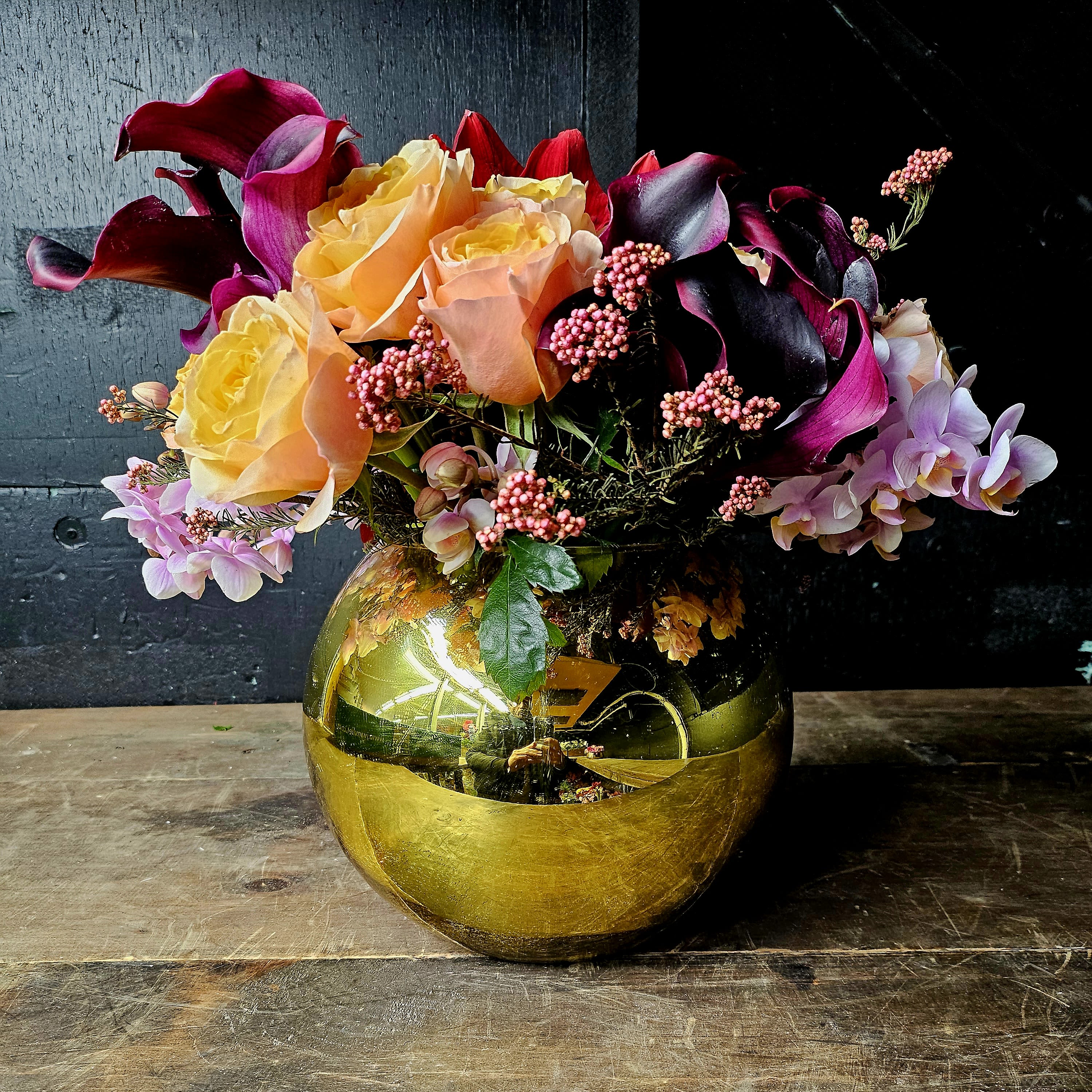 Radiant Elegance: Calla Lilies, Roses, and Orchids in a Golden Vase - Elevate any space with this radiant floral arrangement featuring velvety plum calla lilies, vibrant peach and yellow roses, delicate pink orchids, and soft pink hypericum berries. Presented in an opulent golden vase, this arrangement exudes sophistication and charm, making it the perfect centerpiece for special occasions, celebrations, or as a luxurious gift.  Premium Flowers:  Calla Lilies: Deep plum for an elegant touch.  Roses: Peach and yellow, symbolizing joy and admiration.  Orchids: Soft pink, representing beauty and strength.  Hypericum Berries: Pink accents for texture and charm.   Wrapping: Carefully wrapped for transit and delivered with care to ensure the arrangement arrives in pristine condition.  Occasions: Perfect for anniversaries, birthdays, dinner parties, or any event where you want to make a lasting impression.  Size:  Standard: Approximately 14-16 inches tall.  Deluxe: Taller arrangement with additional blooms.   Care Instructions:  1. Place in a cool, shaded area away from direct sunlight or heat.   2. Add fresh water daily to keep the flowers hydrated.   3. Re-trim stems if needed to ensure optimal water absorption. 