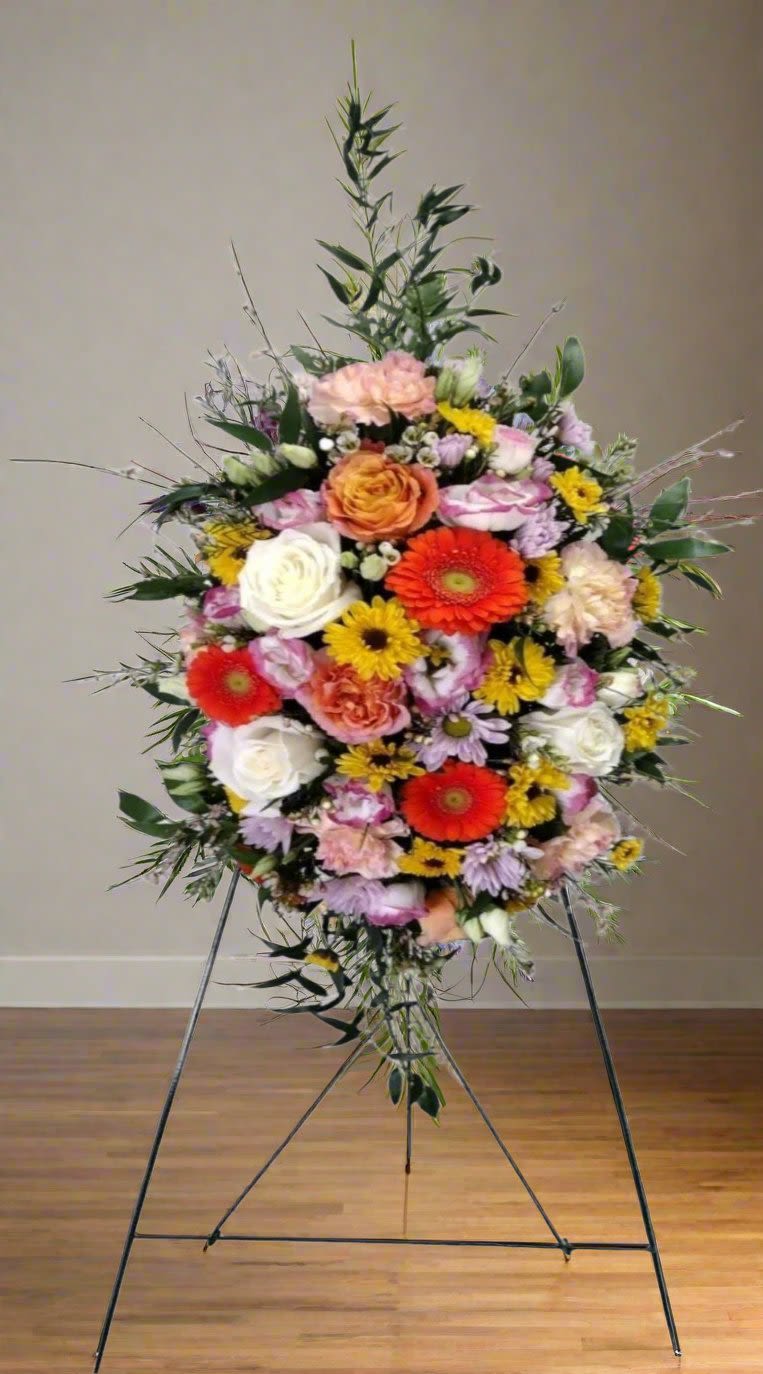 Floral Symphony Easel Spray - The Floral Symphony Easel Spray is a stunning display of vibrant colors, featuring a delightful mix of purples and pinks, accented by lively splashes of orange, white, and yellow. This exquisite arrangement includes an array of blooms such as Gerbera daisies, roses, carnations, lisianthus, viking poms, and wax flowers, all beautifully complemented by lush greens and filler elements.  When you request custom designs, we will strive to create arrangements that closely mirror the image you have in mind. Please remember that each design is one-of-a-kind and tailored specifically for you. Given the natural variability of flowers, some color substitutions may be necessary, and the prices and availability of seasonal blooms can vary.