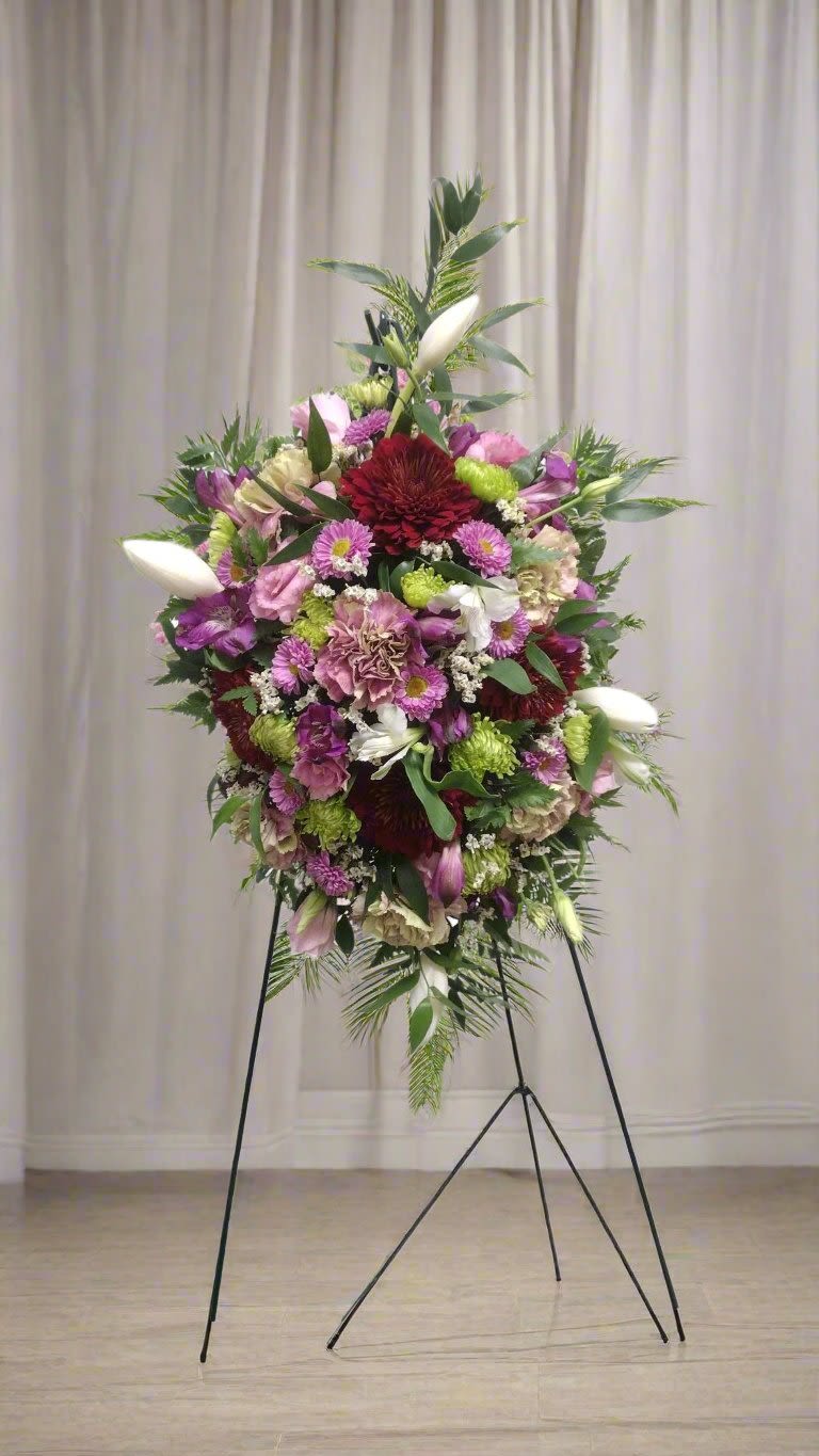 Lavender Harmony Easel - This enchanting easel spray is a stunning composition that beautifully combines shades of lavender, maroon, lime, and white. The arrangement features a charming bouquet made up of tulips, carnations, asters, mums, and stock, presenting a lively showcase of floral elegance. The memorial easel spray is distinguished by its exceptional mix of Matsumoto asters, lisianthus, carnations, alstroemeria, poms, and tulips, all enhanced by rich greenery and filler accents.  When you request custom designs, our goal is to craft arrangements that closely resemble the reference image you provide. It's essential to keep in mind that each design is unique and specifically created for you. Due to the natural variations in flowers, some color adjustments may be required, and the prices and availability of seasonal blooms can fluctuate.