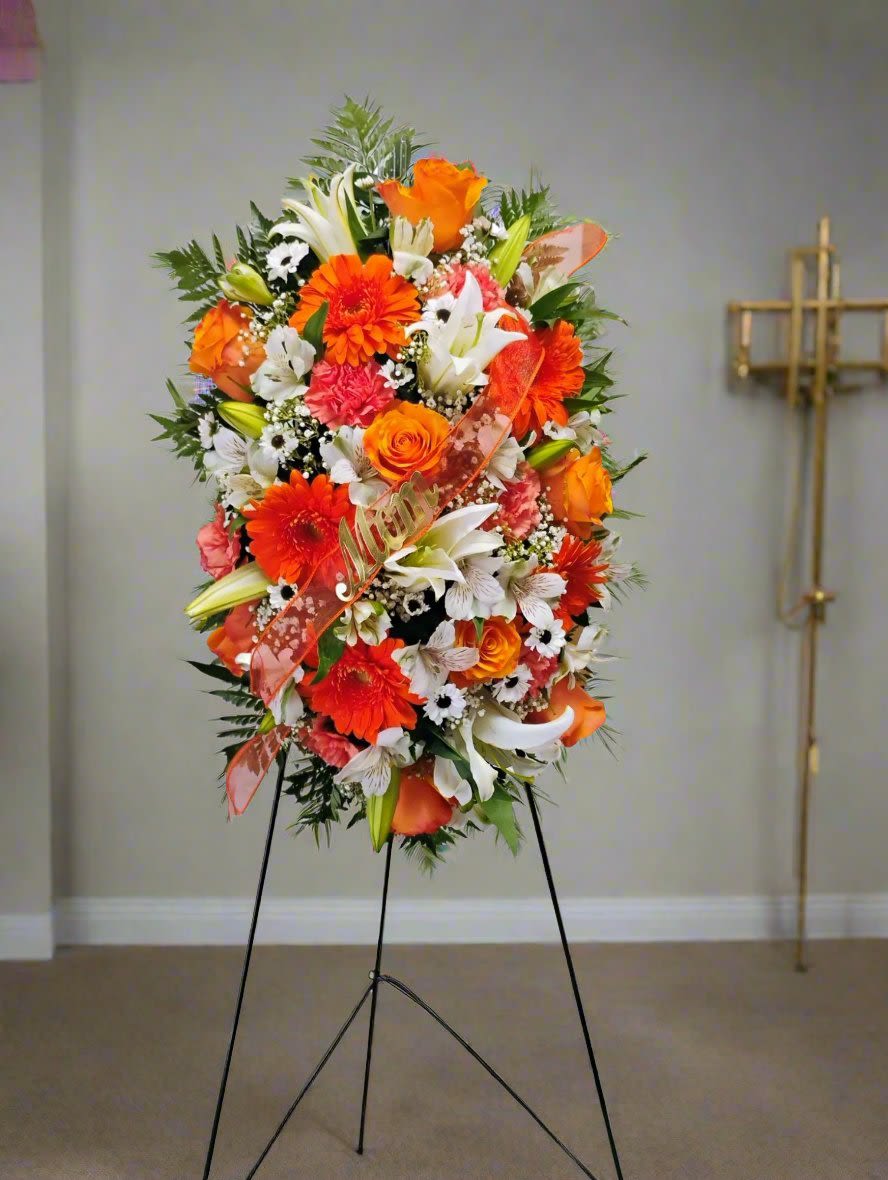 Orange Blossom Serenity Arrangement - Elevate any memorial with our stunning standing floral arrangement, featuring a vibrant blend of orange and white flowers. This exquisite easel spray showcases fresh roses, elegant lilies, cheerful gerbera daisies, and delicate carnations, all beautifully complemented by lush greens and filler flowers.  Perfect for honoring loved ones, this striking color combination is suitable for both men and women, making it a versatile choice for any occasion. Choose this breathtaking arrangement to convey your heartfelt sentiments and celebrate a life well-lived.