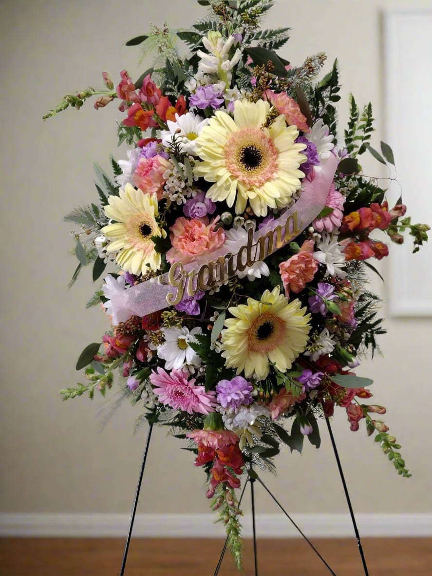Pastel Parting Floral Trubute - A breathtaking easel spray featuring fresh flowers in delicate pastel tones. This lovely arrangement includes gerbera daisies, carnations, snapdragons, and poms, beautifully intertwined to form a heartfelt tribute to celebrate your loved one. You can request a personalized script in the notes if desired.  For custom designs, we will work diligently to replicate the image you share with us. Please note that every creation is unique and tailored just for you. Given the natural differences in flowers, some color changes might be necessary. Additionally, prices and availability of seasonal blooms can vary.&nbsp;