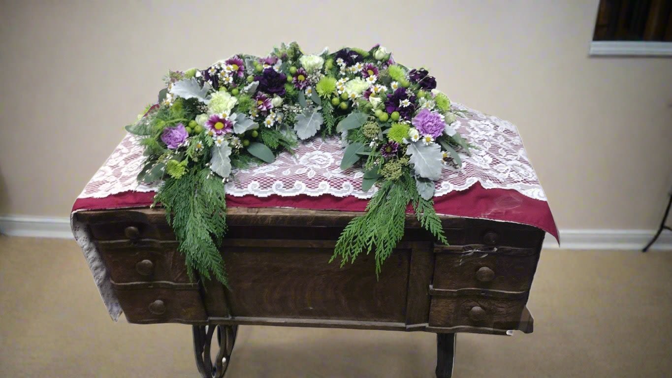 Purple &amp; Green Natural Memorial - Elevate your memorial tribute with our stunning floral arrangement, featuring rich purple and green shades that beautifully complement the natural green backdrop. This heavy piece is thoughtfully crafted with vibrant carnations, cheerful daisies, elegant poms, and unique hypericum berries, all nestled among lush mixed greens. Personalize your tribute with an optional ribbon and script, making it a heartfelt expression of love and remembrance. Choose this exquisite arrangement to honor your loved one in a truly memorable way.  **When you request custom designs, we will create them to closely resemble the picture. Keep in mind that each design is unique and made just for you. Due to the nature of flowers, color substitutions may be required. Prices and availability of seasonal flowers can also differ.