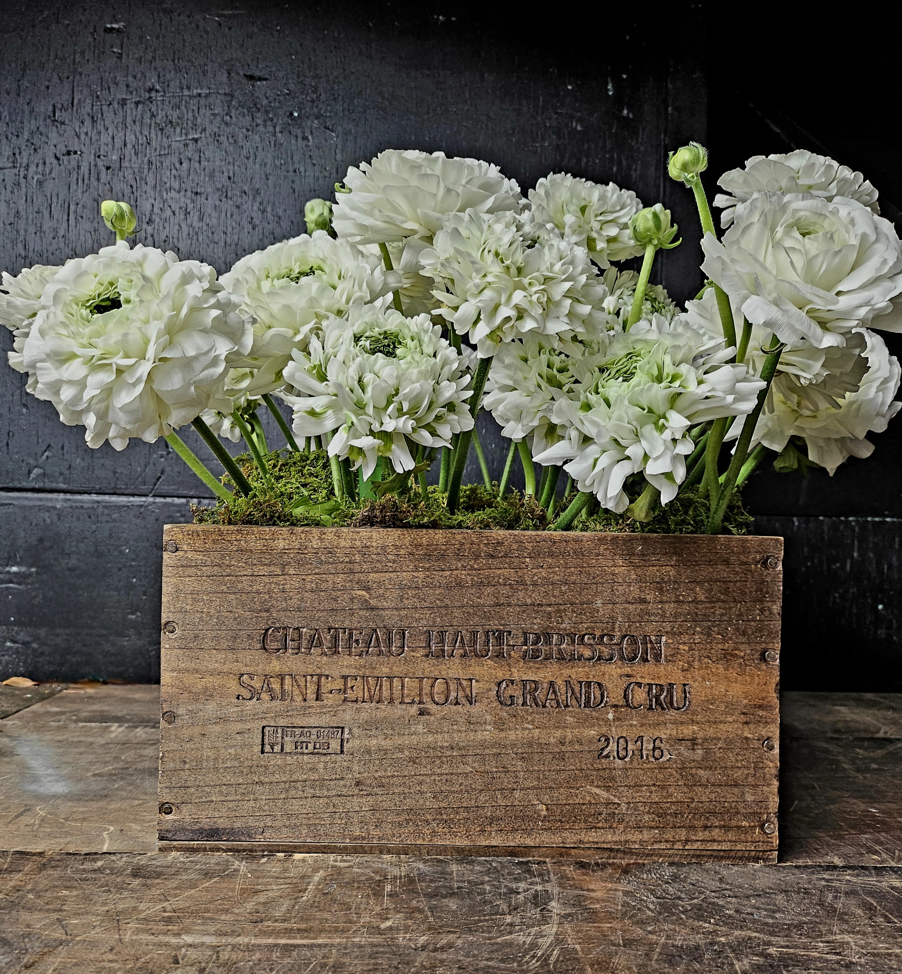 Rustic Elegance: White Ranunculus in Reclaimed Wooden Box - Experience timeless beauty with this charming arrangement of premium white ranunculus artistically nestled in a reclaimed wooden wine box. Accentuated with lush green moss, this design exudes rustic elegance, perfect for adding a sophisticated touch to any room. The delicate layers of ranunculus petals symbolize purity and grace, making this arrangement ideal for weddings, anniversaries, or simply celebrating life's special moments.  The wooden box, engraved with a vintage wine label, lends an earthy, nostalgic vibe that complements the soft, fresh blooms. Designed with care and attention to detail, this piece is a statement of refined simplicity.  Details:  Flowers Included: White Ranunculus, Green Moss.  Container: Reclaimed wooden wine box with engraved details.  Size: Approx. 12” (L) x 6” (W) x 9” (H).  Premium Quality: Each bloom is hand-selected to ensure freshness and longevity.   Occasions:  Perfect for weddings, anniversaries, housewarming gifts, or as an elegant centerpiece.   Care Instructions:  Place in a cool location away from direct sunlight and heat.  Mist moss lightly to retain freshness.  No additional watering required due to the moss base.  Let this unique arrangement bring a touch of rustic sophistication to your next event or as a thoughtful gift for someone special! 