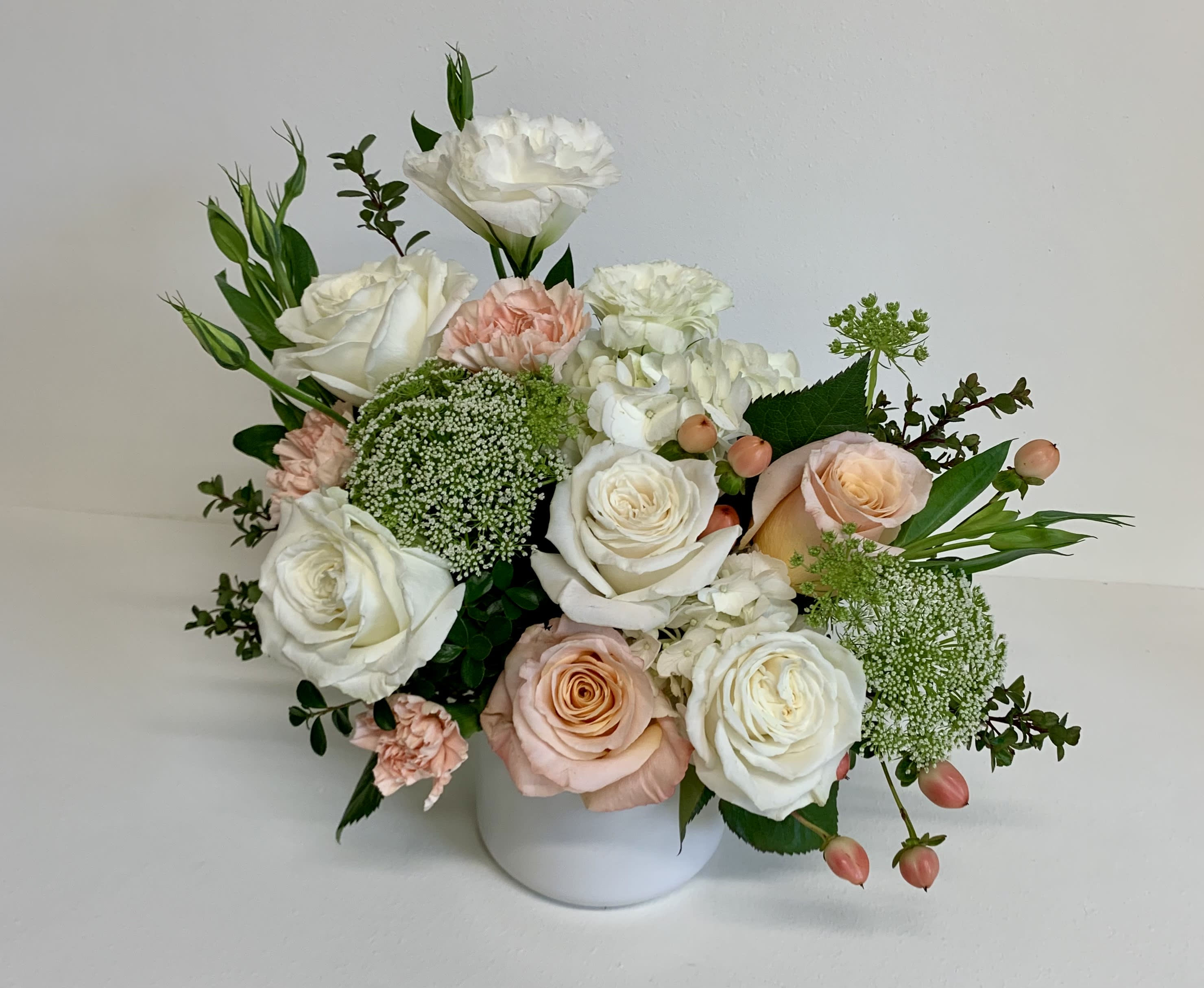 Cambria - This arrangement features playa blanca and shimmer roses, peach carnations, white lisianthus, peach hypericum, queen anne's lace and a white hydrangea with a touch of box wood and delivered in a white ceramic vessel.