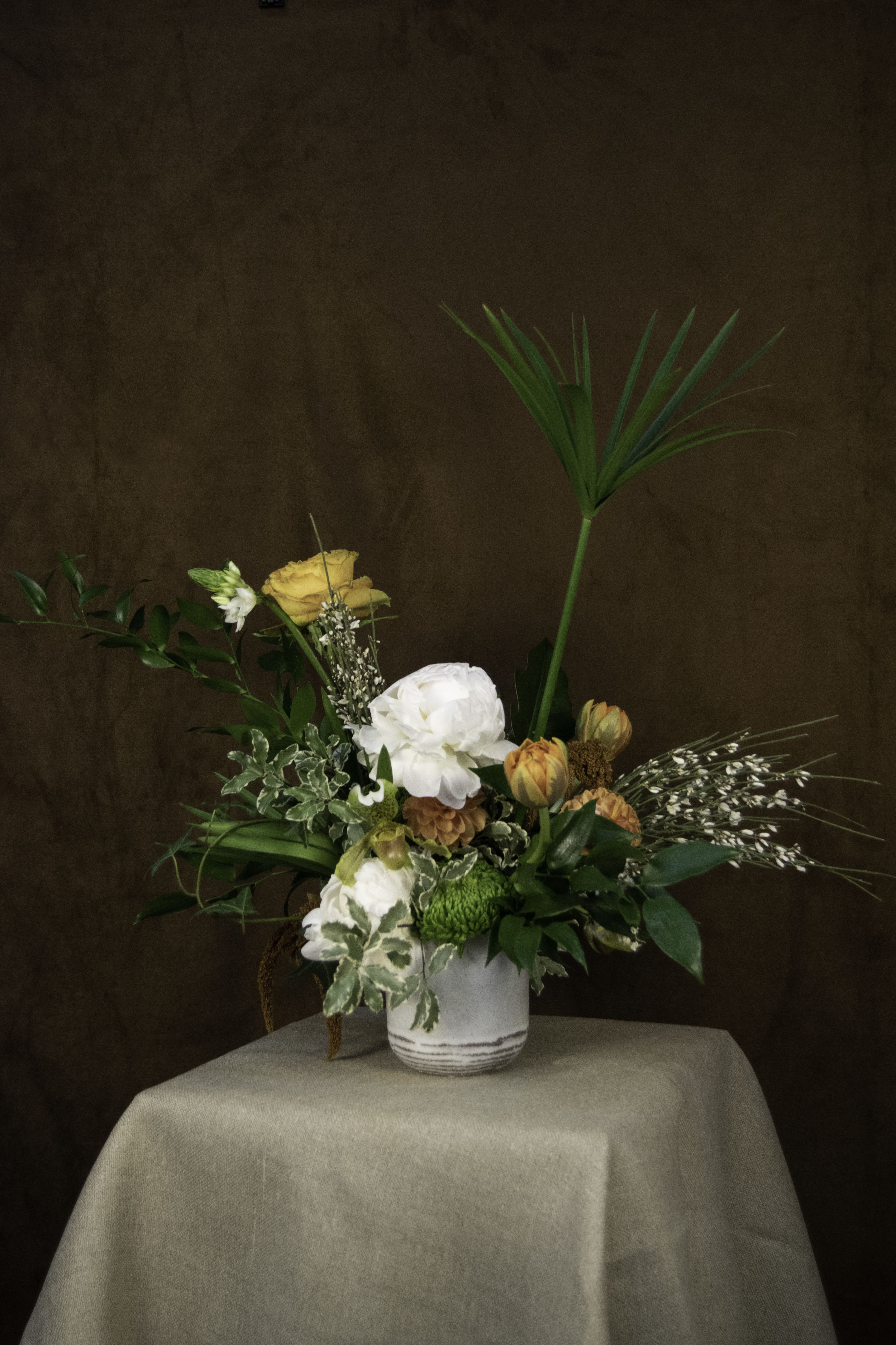 Pansodan - The &quot;Pansodan&quot; floral arrangement by The Burmese Cat studio radiates natural charm and balanced elegance. Anchored by a pristine white peony, it features warm orange tulips, golden-hued roses, and bursts of green chrysanthemums. Delicate sprays of white buds and textured greenery, including lush leaves and variegated ivy, add layers of depth. A tall palm leaf rises gracefully, evoking a sense of vertical drama.  Set in a simple white vase, this arrangement captures the timeless sophistication and cultural richness of its namesake, Pansodan.  ---------------------------------------------------  At The Burmese Cat Floral Studio, we celebrate the uniqueness of every flower and vase. While the photo captures the spirit of your arrangement, some elements—like one-of-a-kind vessels or seasonal stems—might not always be exactly the same. However, each arrangement will embody the same charm, elegance, and artistic flair.  If substitutions are needed due to weather, seasonal shifts, or market availability, we’ll stay true to the mood, colors, and style you’ve chosen. Rest assured, any replacements will match or exceed the value of the original design.  If there’s something specific that’s a must-have for your arrangement, just let us know in the notes at checkout or reach out directly—we’ll do our best to make it happen!  ---------------------------------------------------