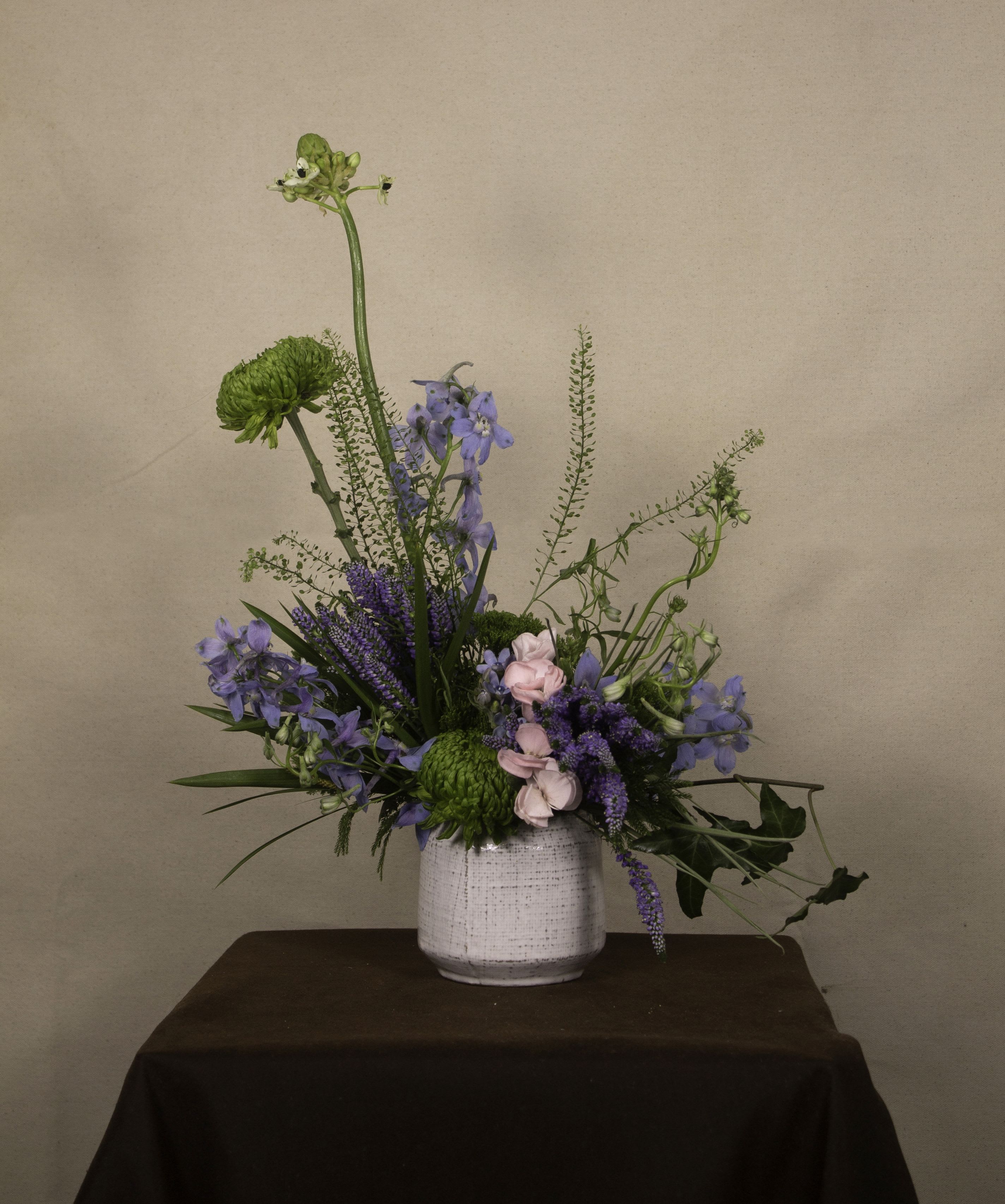 Dagon - The &quot;Dagon&quot; floral arrangement features a mix of green chrysanthemums, blue delphiniums, and soft pink roses. Accents of lavender veronica and flowing greenery add texture and height, while a white ceramic vase keeps the design clean and simple. This arrangement offers a fresh and calming look, perfect for any space. ---------------------------------------------------  At The Burmese Cat Floral Studio, we celebrate the uniqueness of every flower and vase. While the photo captures the spirit of your arrangement, some elements—like one-of-a-kind vessels or seasonal stems—might not always be exactly the same. However, each arrangement will embody the same charm, elegance, and artistic flair.  If substitutions are needed due to weather, seasonal shifts, or market availability, we’ll stay true to the mood, colors, and style you’ve chosen. Rest assured, any replacements will match or exceed the value of the original design.  If there’s something specific that’s a must-have for your arrangement, just let us know in the notes at checkout or reach out directly—we’ll do our best to make it happen!  ---------------------------------------------------