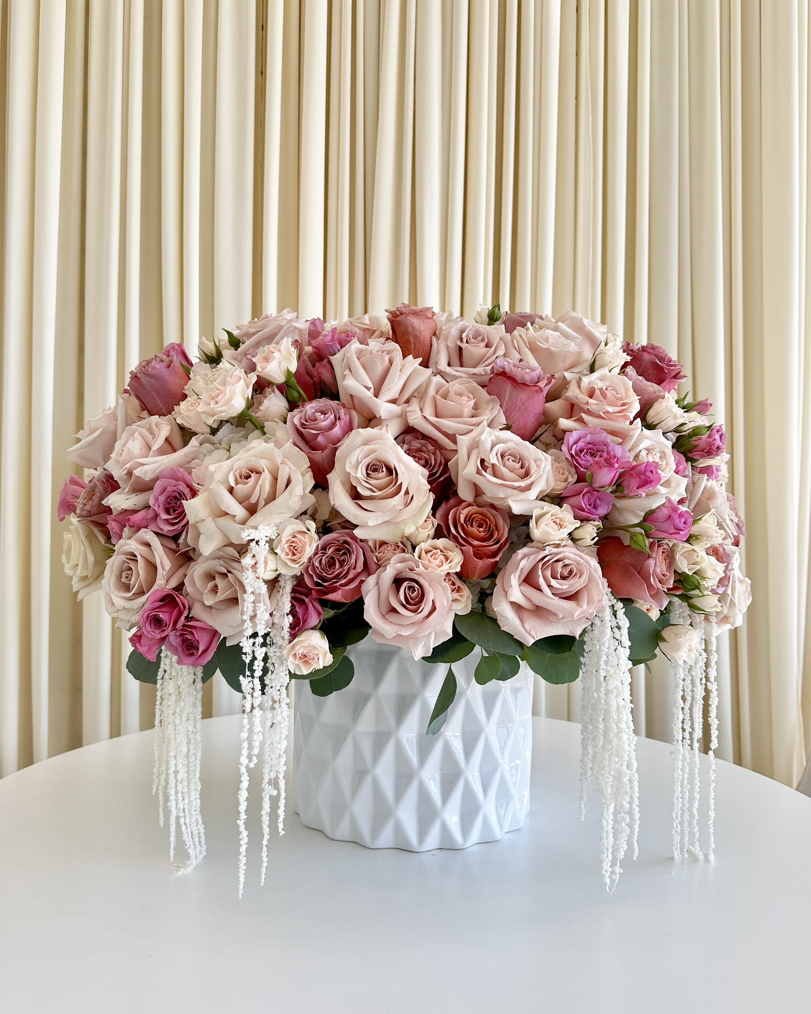 Blushing Rose Cascade - Introducing the &quot;Blushing Rose Cascade,&quot; a spectacular floral arrangement that captures the heart with its radiant shades of pink roses and spray roses. This enchanting bouquet is a true masterpiece, perfect for expressing your deepest emotions or adding a touch of elegance to any space.  The &quot;Blushing Rose Cascade&quot; is a symphony of pink hues, from soft pastels to vibrant blooms, all carefully selected to create a harmonious blend of colors and textures. Each rose is a testament to nature's beauty, with petals that unfurl like delicate poetry.  What sets this arrangement apart is the inclusion of dried amaranthine, gracefully draping from the pristine white vase. This unexpected element adds a touch of whimsy and ethereal charm, creating a mesmerizing visual display that evokes a sense of wonder.  Whether you're celebrating a special occasion, expressing your love, or simply brightening someone's day, the &quot;Blushing Rose Cascade&quot; is the ideal choice. Its timeless beauty and romantic allure will captivate hearts and leave a lasting impression.  Bring a touch of enchantment into your life or surprise a loved one with this remarkable creation from our flower shop. Order the &quot;Blushing Rose Cascade&quot; today and let its beauty fill your world with joy and admiration.