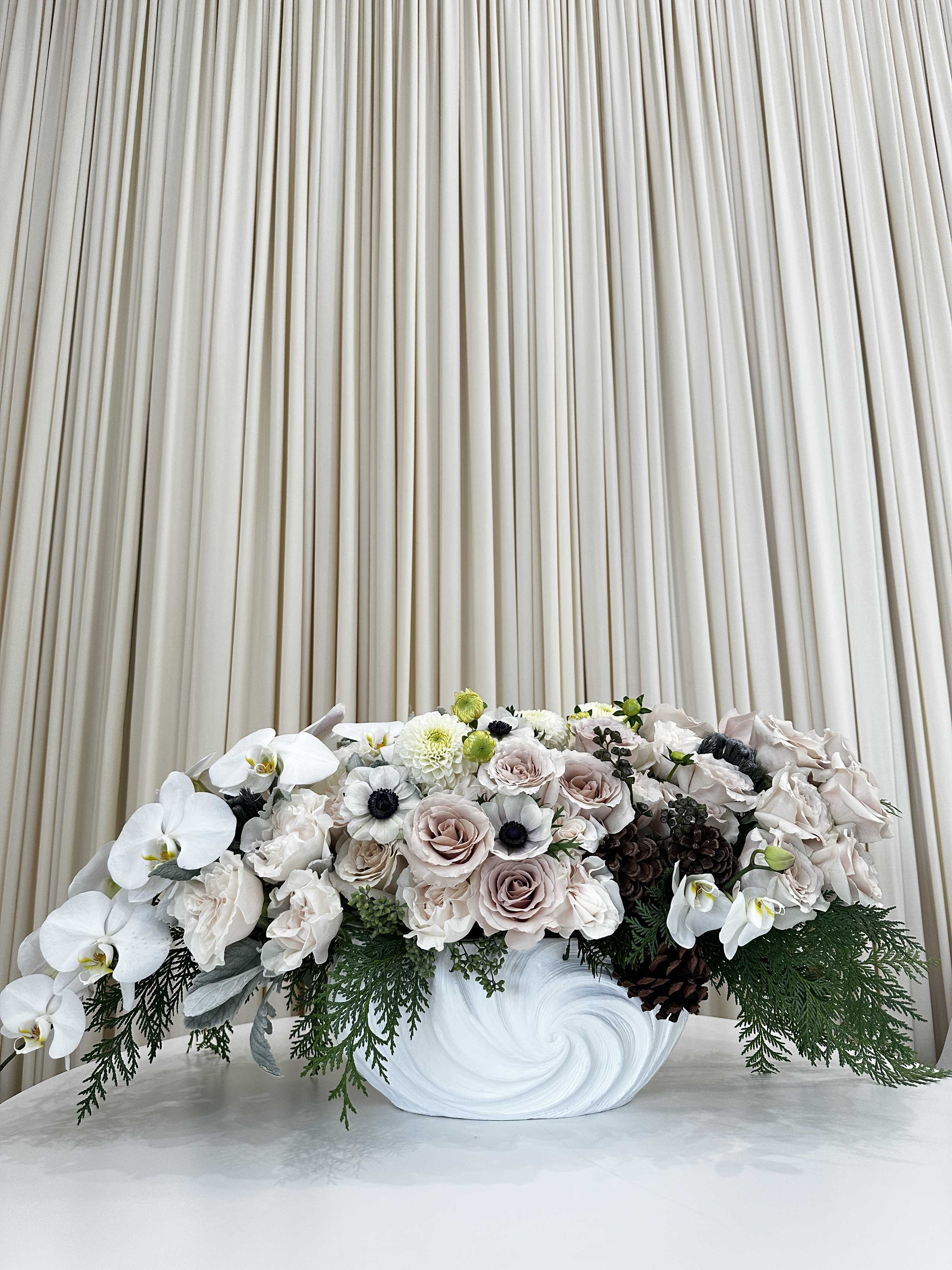 Winter Whisper - Embrace the enchantment of winter with this soft and elegant floral arrangement. Featuring an assortment of white roses, hydrangeas, and orchids, accented with pine cones and greenery, this arrangement is sure to bring a touch of peace and serenity to your home.