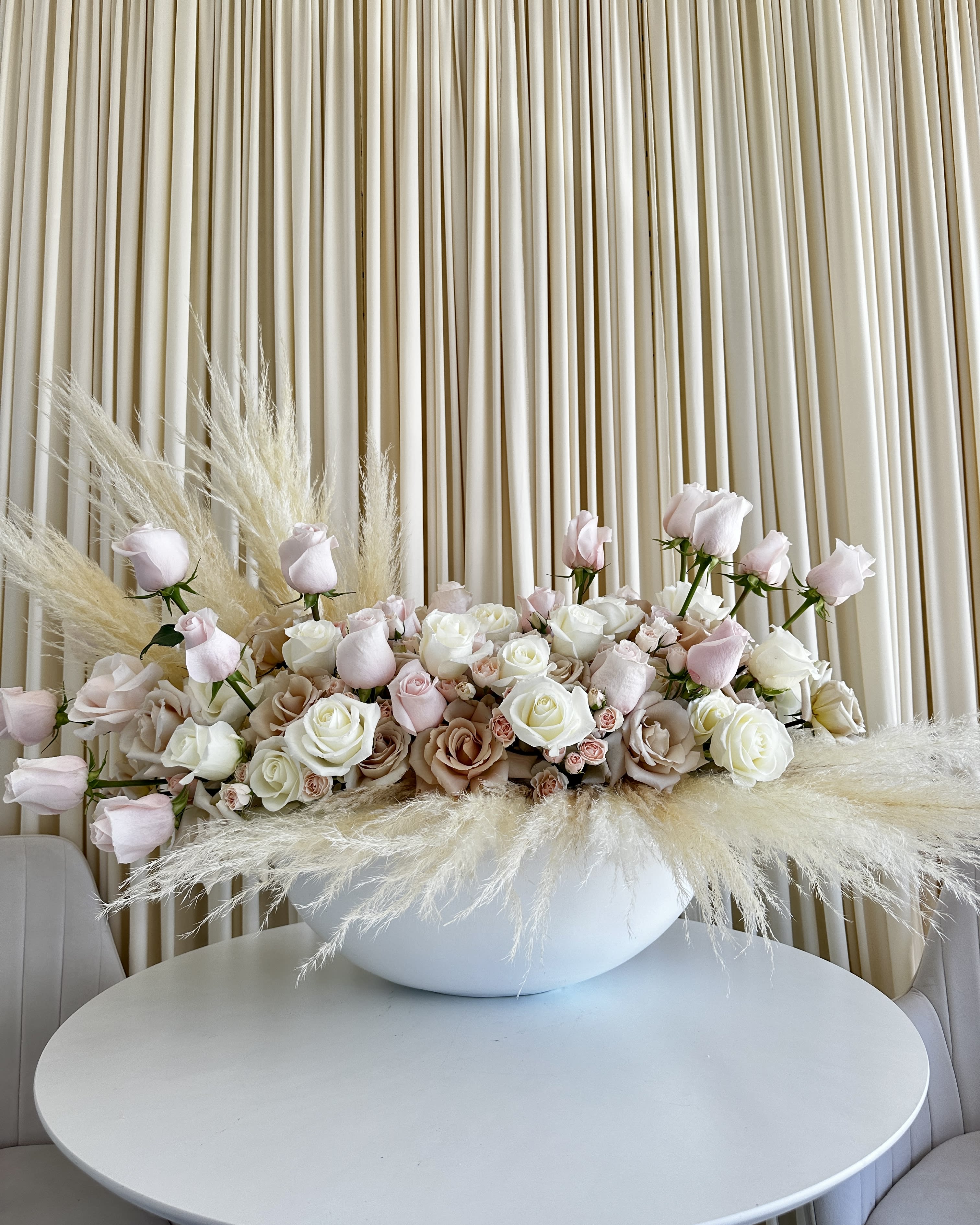 Elegant Elysium Oasis - Welcome to a realm of floral sophistication with our &quot;Elegant Elysium Rose &amp; Pampas Oasis&quot; arrangement. This exquisite creation captures the essence of timeless beauty through a harmonious blend of neutral-colored roses and the graceful allure of pampas grass, all cradled within a pristine white ceramic vase.  Each element of this arrangement has been meticulously curated to convey elegance and purity. The neutral-hued roses, a symbol of grace and admiration, are the centerpiece of this masterpiece. Their delicate petals, ranging from creamy whites to soft blush tones, exude a sense of understated luxury.  Surrounding the roses, the ethereal pampas grass creates an enchanting sense of movement and lightness. The feathery plumes, like whispers of nature, add a touch of wild elegance to the composition. Pampas grass symbolizes freedom and eternal love, making it a meaningful addition to any floral arrangement.  Nestled within a pristine white ceramic vase, this arrangement exudes a sense of refinement and timelessness. The clean lines and pure color of the vase enhance the overall aesthetic, making it suitable for any interior décor style.  Whether you're seeking the perfect gift to express your feelings or looking to elevate your living space, our &quot;Elegant Elysium Rose &amp; Pampas Oasis&quot; is the embodiment of natural beauty and elegance. Let this arrangement bring a touch of tranquility and sophistication into your life or delight a loved one with a gift that speaks volumes without uttering a word. Experience the allure of neutral beauty with this breathtaking floral masterpiece.