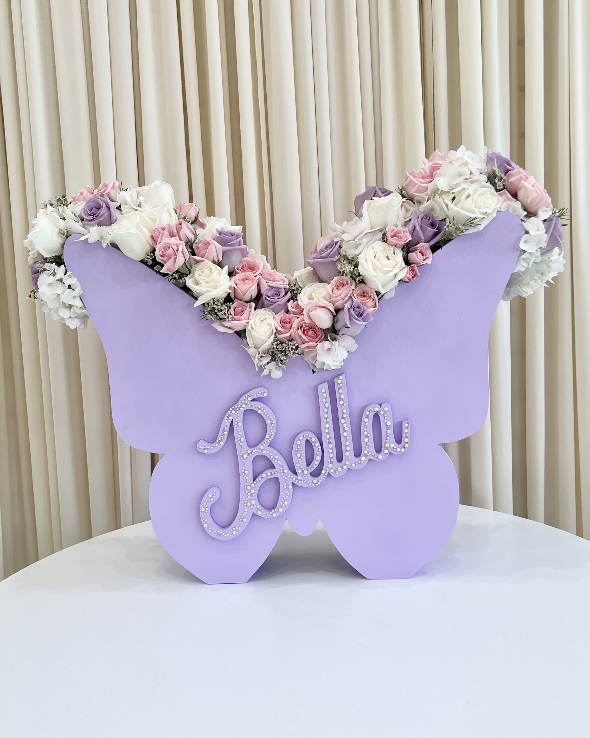 Fluttering Butterfly - Embrace the enchantment of &quot;Fluttering Butterfly,&quot; a custom butterfly-shaped vase filled with an array of elegant white, pink, and purple florals. This exquisite arrangement exudes a delicate charm, gracefully capturing the beauty of nature's palette.  Each blossom, carefully selected for its pristine beauty, dances in harmony within the vase, bringing a symphony of colors and scents. The soft whites signify purity and tenderness, the gentle pinks symbolize admiration and affection, while the gentlr purples evoke a sense of wonder.  Personalize the experience with a customized name elegantly inscribed on the vase, making it a cherished keepsake to treasure for years to come. Perfect as a unique gift for loved ones, this butterfly vase arrangement will flutter its way into hearts, leaving a lasting impression of beauty and thoughtfulness. 