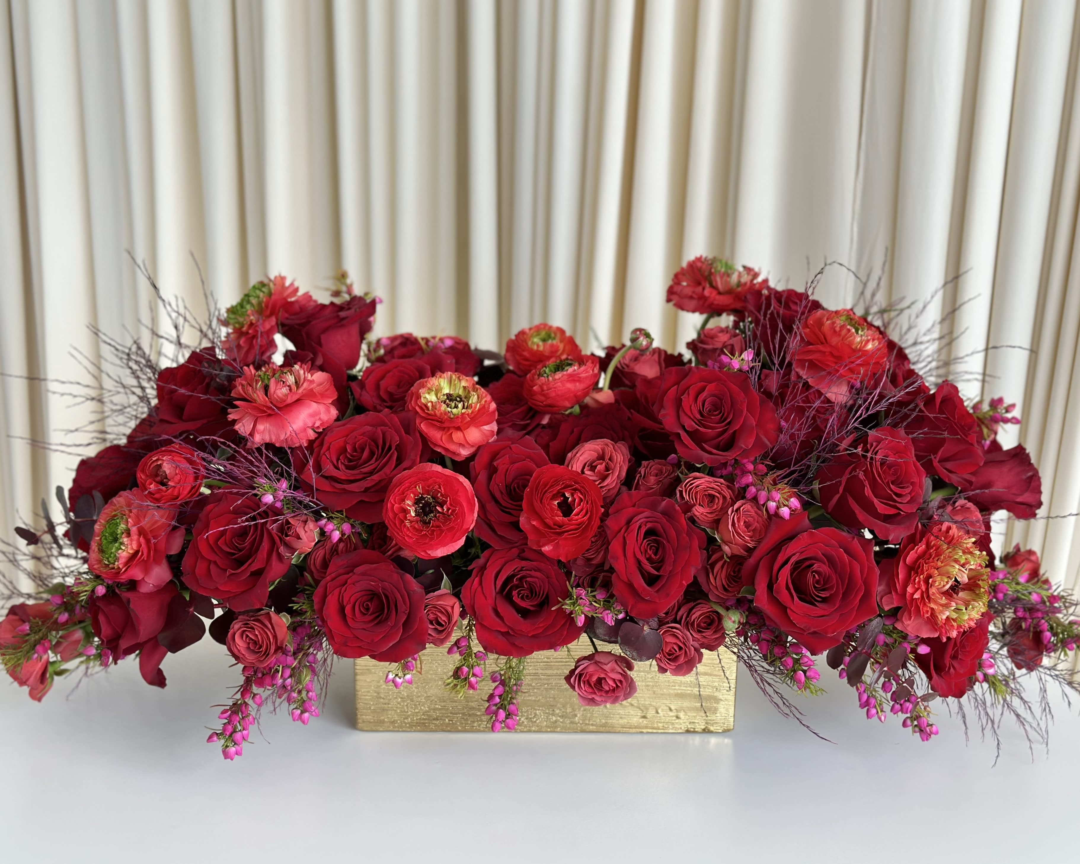 Crimson Radiance - Behold the captivating allure of &quot;Crimson Radiance,&quot; an exquisite flower arrangement that exudes passion and charm. Nestled within a luxurious gold vase, this masterpiece combines the timeless beauty of all-red roses and red ranunculus flowers, accented by delicate twigs and pops of vibrant fuchsia blooms.  The velvety red roses symbolize deep love and affection, while the enchanting red ranunculus flowers add a touch of delicacy and grace. Together, they create a symphony of red hues, igniting the senses and expressing profound emotions.  Intertwined with natural twigs for an earthy element and complemented by fuchsia flowers that exude playfulness and energy, &quot;Crimson Radiance&quot; becomes an exquisite fusion of passionate romance and modern sophistication.