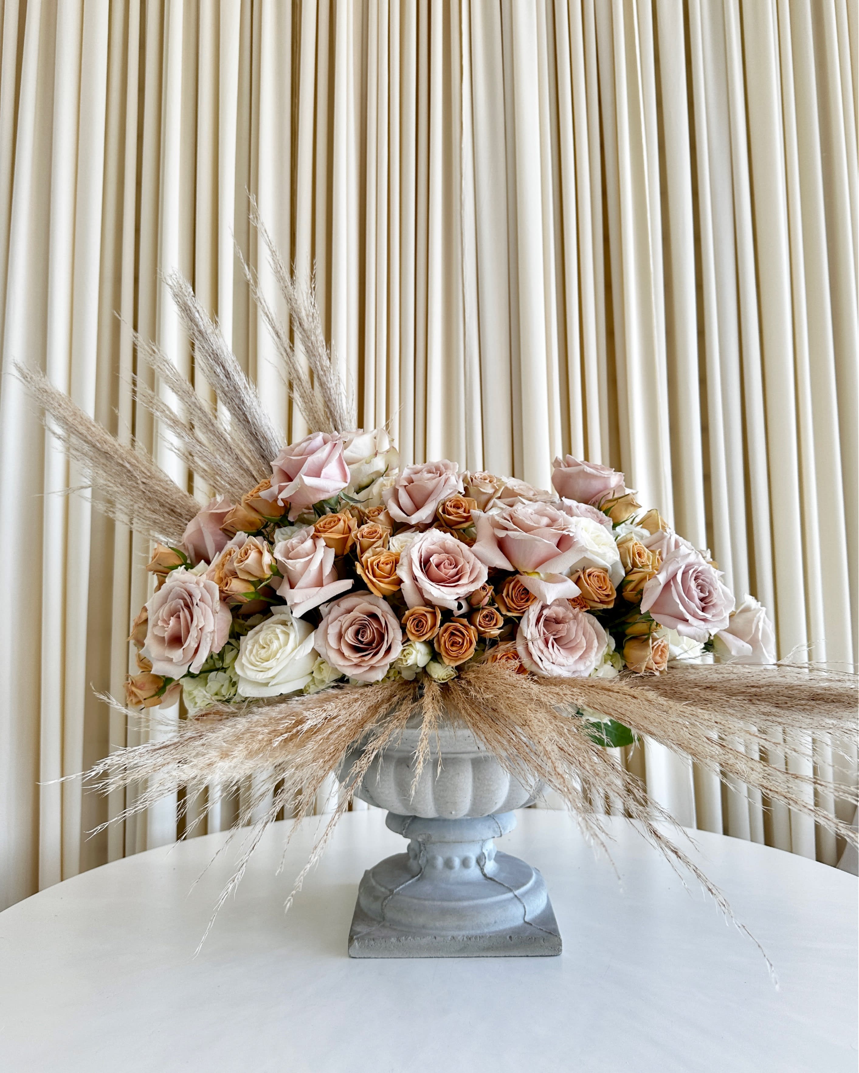 Elegance in Bloom Pedestal Arrangement - Introducing our exquisite &quot;Elegance in Bloom Pedestal Arrangement&quot; – a masterpiece of floral artistry that will add a touch of sophistication and natural beauty to any space. This stunning arrangement combines the timeless charm of pink, white, and rusty orange roses, expertly arranged in a graceful pedestal vase, and elegantly framed by the soft, feathery plumes of lush pampas grass.  Each element of this arrangement has been carefully selected to create a harmonious and captivating composition. The soft, delicate petals of the roses provide a romantic allure, while the warm hues of rust and delicate pinks add a playful touch. The pristine white roses symbolize purity and grace, making this arrangement perfect for a variety of occasions, from weddings to anniversary celebrations.  The star of this arrangement, the pampas grass, adds a unique and trendy twist to the traditional floral display. Its feathery plumes evoke a sense of tranquility and freedom, as if capturing the essence of a meadow at sunrise.  Whether you're looking to impress a loved one, elevate your home decor, or make a statement at a special event, our &quot;Elegance in Bloom Pedestal Arrangement&quot; is the ideal choice. Let the beauty of these meticulously curated blooms and the elegance of the pedestal vase bring nature's grace and charm into your life.